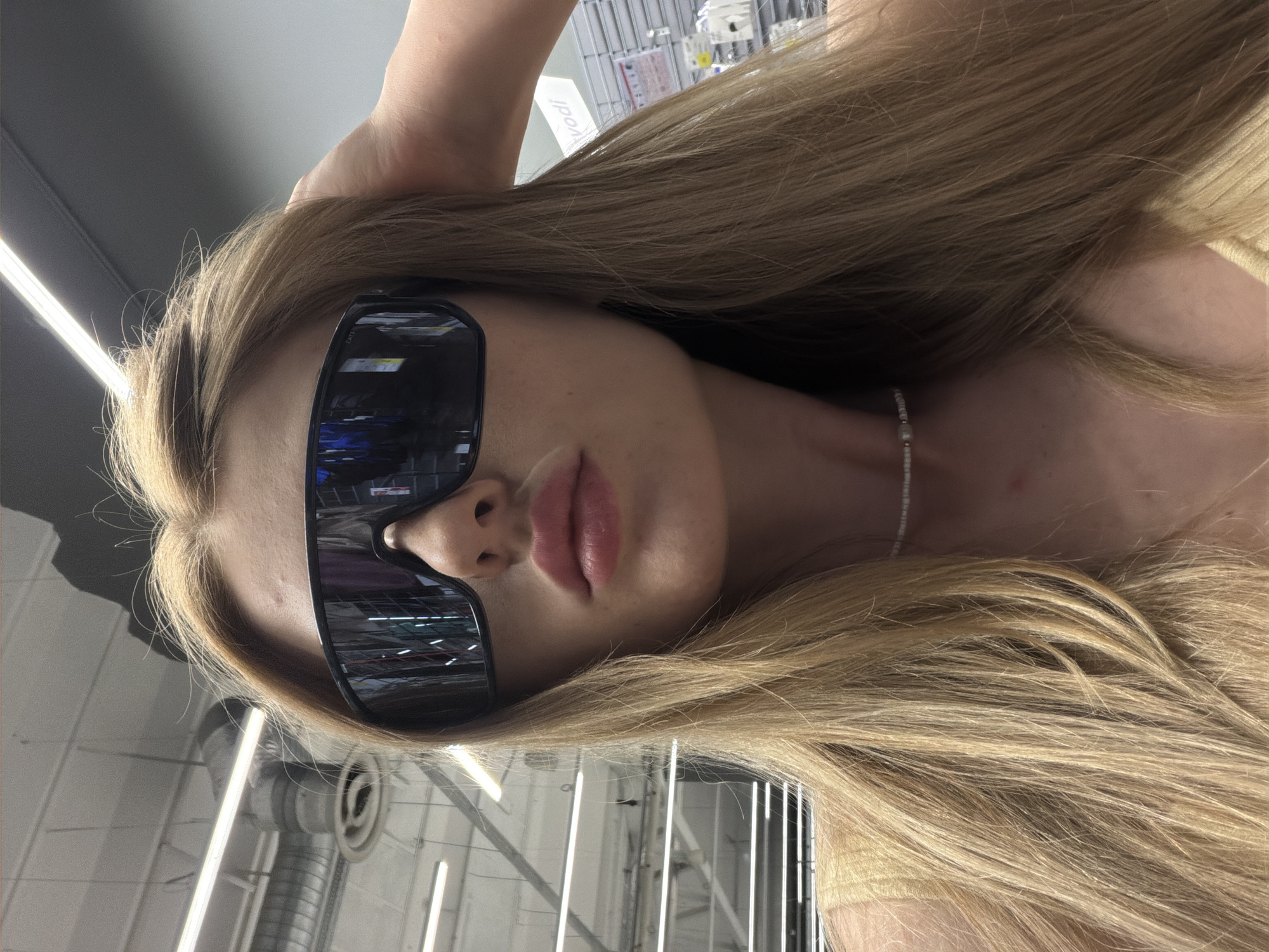 A young woman with long blonde hair, wearing large dark sunglasses and a thin beaded necklace, poses for a selfie. She is outdoors in what appears to be a retail environment, with overhead fluorescent lighting, shelving units, and ventilation systems visible in the background. The woman's lips are pursed, and her right hand is raised, touching her hair. She is wearing a light-colored, possibly yellow, top.