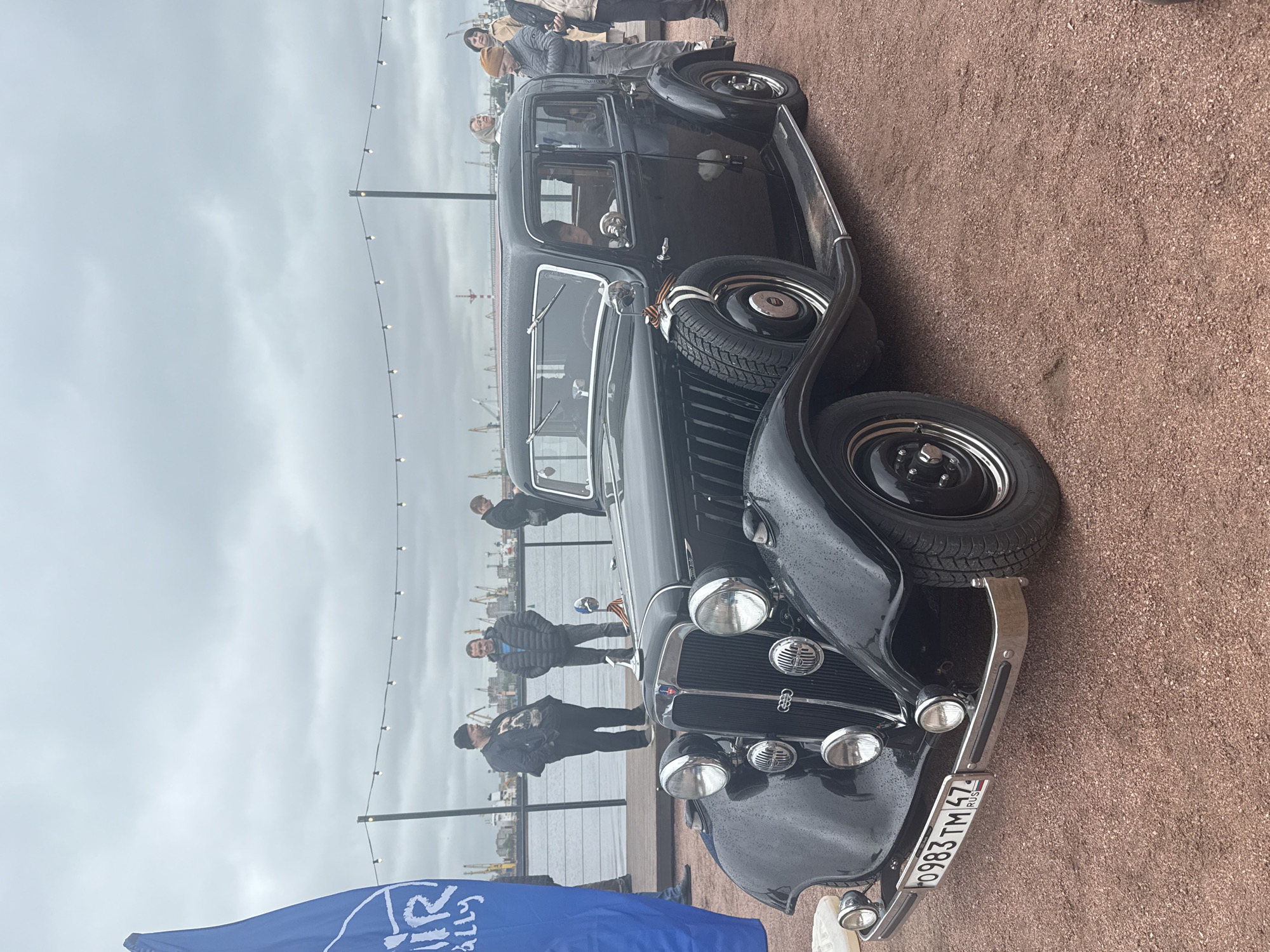 The image depicts a black classic car parked alongside a waterfront in Saint Petersburg, Russia. The car is the main subject, with a license plate reading "O983 TM 47 RUS". A spare tire is mounted on the side of the car, just behind the front wheel. People are standing near a railing overlooking the water. The background includes the city skyline, a body of water, and an overcast sky. The ground is covered with gravel. A blue banner with white text is visible in the bottom left corner. Strings of lights are strung between poles behind the group of people.