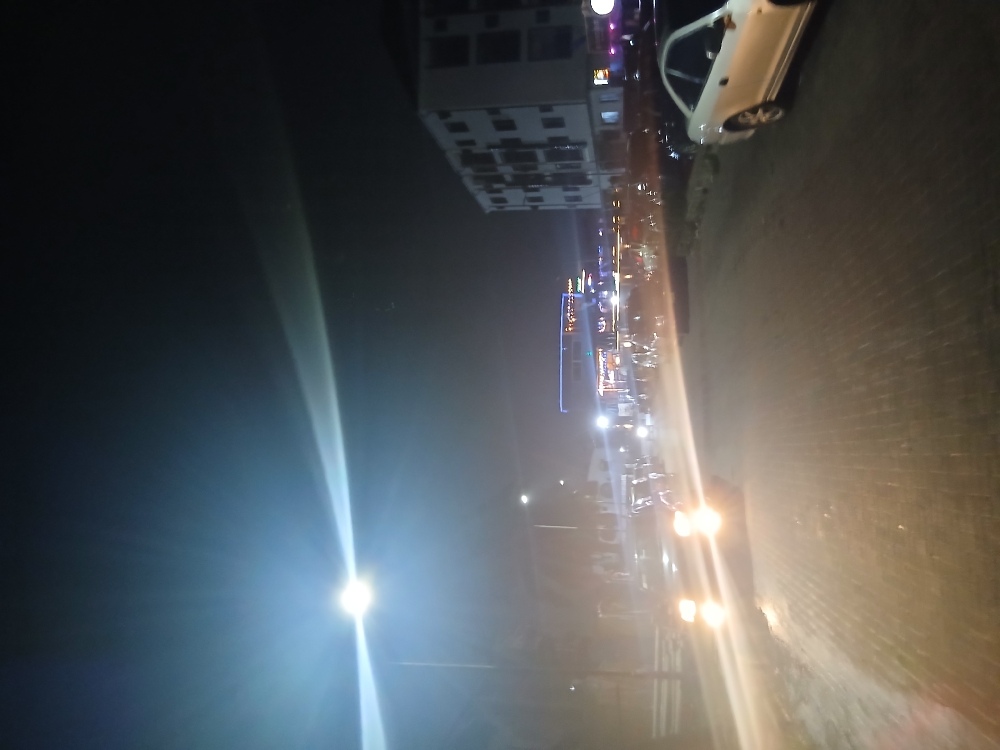 The image depicts a nighttime street scene in Lagos, Nigeria. A white car is parked on the right side of the frame, near a brick or paved road. Buildings and streetlights line the road. There is a visible streetlight. Other vehicles and pedestrians are present, though details are obscured by the darkness and distance. Overall, the scene portrays a typical urban environment at night.