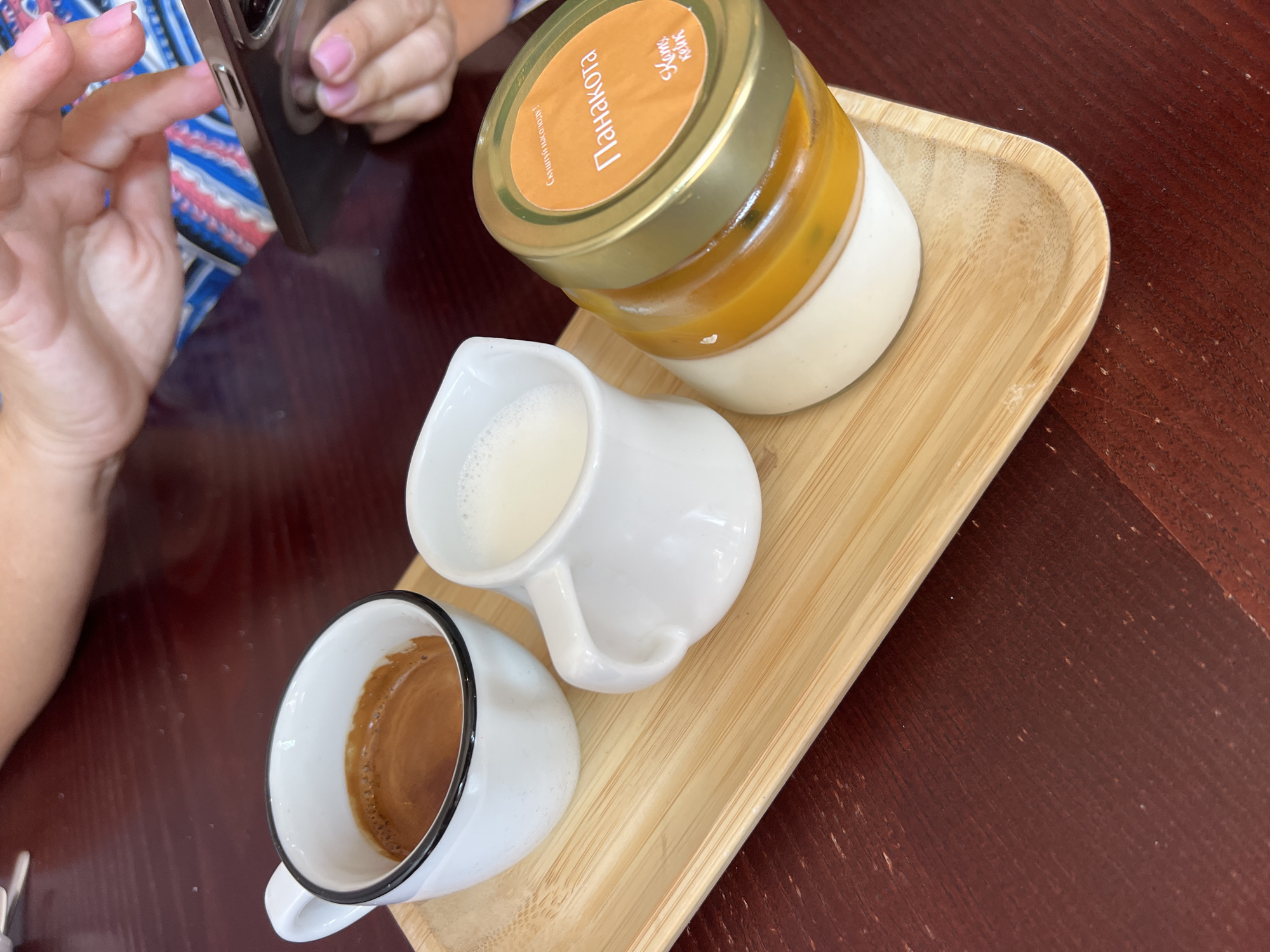 An overhead medium shot captures a wooden tray on a dark red table, presenting a coffee and dessert set. A white ceramic cup with a black rim holds a dark brown espresso. Next to it, a small white pitcher contains frothy white milk. To the right, a glass jar with a gold lid contains a layered dessert, with the top layer being a vibrant orange hue, likely a fruit jelly or passion fruit topping, over a creamy white base. An orange label on the jar displays Cyrillic text, reading "ПАНАКОТА" (Panna Cotta) prominently, with smaller text above and below. The lower right portion of the image reveals the corner of a person's hand, with pink-painted fingernails, holding a dark-colored smartphone. In the background, another person's hands are visible, also interacting with the phone. The setting suggests a cafe or restaurant environment in Fontanka, Ukraine.