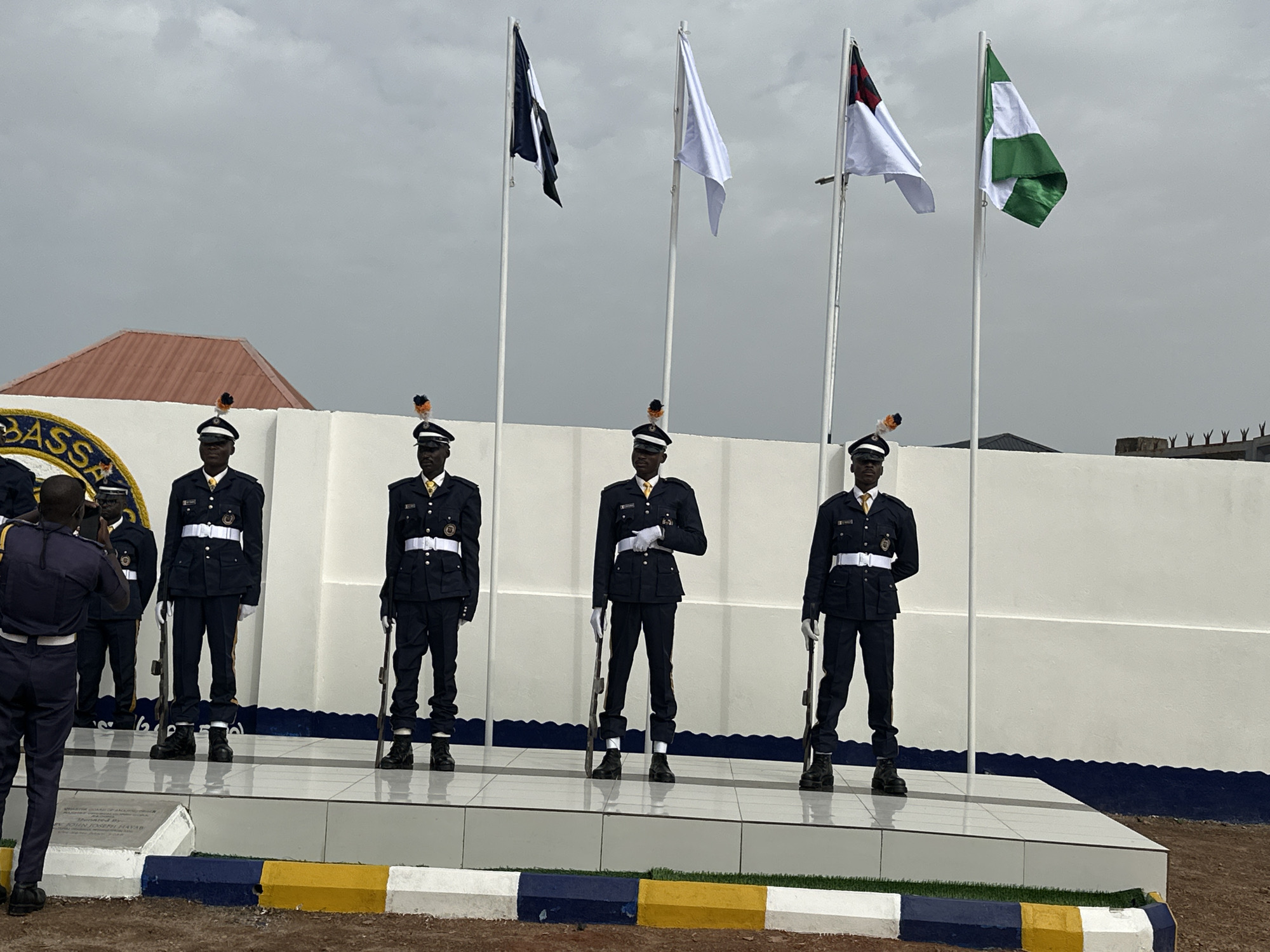 The image shows five uniformed men standing on a tiled platform in front of a white wall, near Goni Gara, Nigeria. Each man, wearing a dark blue uniform with a white belt and a black hat with an orange plume, holds a rifle. Four flags are displayed behind them on poles: a black flag, a white flag, a red and white flag, and a green and white flag. Another uniformed man stands to the left of the group. The surrounding ground is reddish-brown. The sky is overcast. The roof of a building is visible behind the wall.
