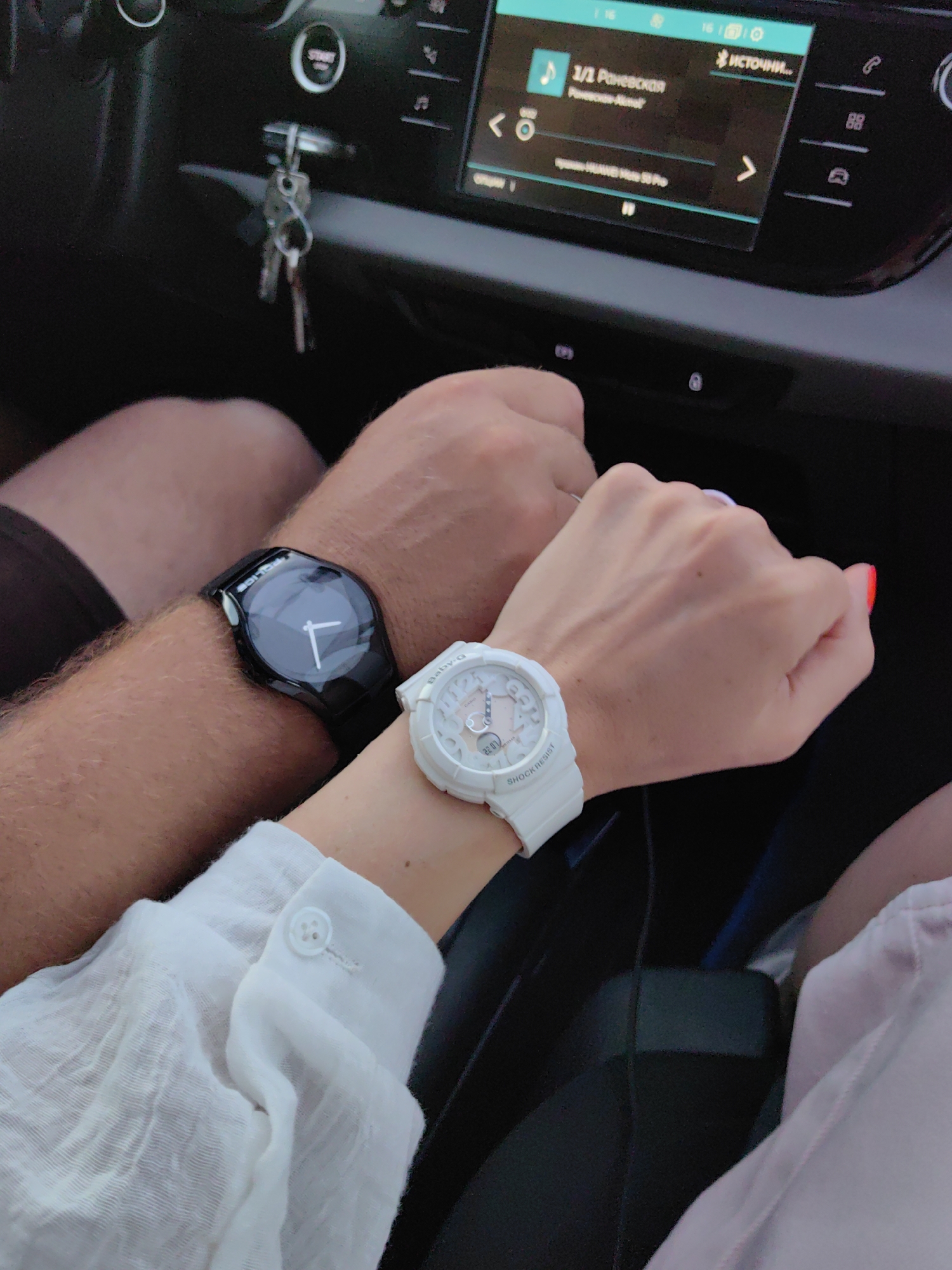 Two individuals are seated in a car, their hands clasped. The person on the left wears a black digital watch, while the person on the right wears a white analog watch and has bright red nail polish. The car's dashboard is visible, featuring a media display that shows music information, and a set of keys hangs from the ignition. The lighting suggests it is daytime. The specific location within Дзяржынскі сельскі Савет, Belarus, cannot be determined from the visual information provided.