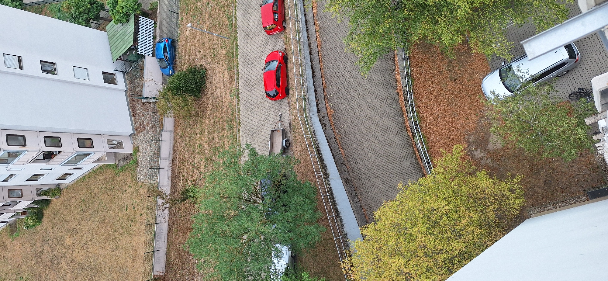 The image, taken from an elevated perspective, shows an outdoor scene in a residential area. In the foreground, a light-colored building facade is visible on the left. A paved area with fallen brown leaves is present, with several trees featuring green and yellow foliage scattered throughout. A silver station wagon is parked on the paved area, alongside a bicycle.
Further back, a road or path paved with interlocking concrete blocks runs horizontally. On this path, two red cars are parked, one behind the other. A small, empty utility trailer is also parked on the paved area, to the left of the red cars. A large tree with green foliage dominates the middle ground on the left side.
In the background, a modern apartment building with multiple balconies and windows is visible. A blue hatchback car is parked on a paved driveway to the right of the building, near a small green-roofed structure with white fencing. Beyond the buildings and paved areas, dry, yellowish-brown grass covers the ground, bordered by a fence.
This scene is located in Karlsruhe, Germany, specifically in the area of Theodor-Heuss-Allee 58, 76135 Karlsruhe, based on the provided context. The overall impression is one of a calm residential street on an autumn day.
