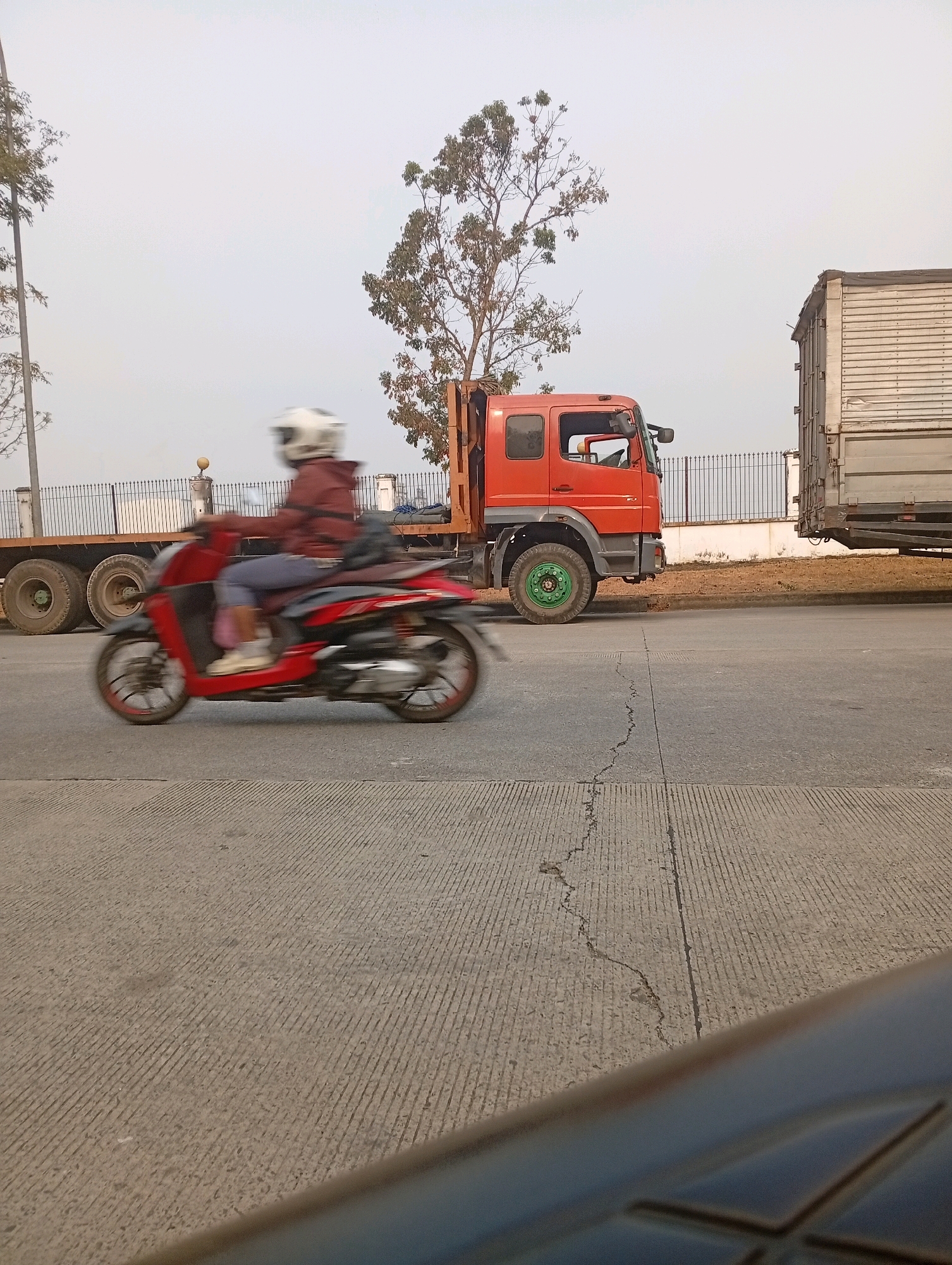 In the city of Sukamahi, Indonesia, a red scooter with a rider wearing a white helmet and a maroon jacket is captured in motion on a paved road. The rider's body and the scooter are blurred due to movement. Behind the scooter, an orange truck is parked, its cab visible. Further in the background, another truck with a white cargo container is parked, and a black metal fence runs along the roadside. Trees with sparse foliage are present near the trucks. The sky is overcast and light in color. The road surface is concrete with parallel textured lines and a visible crack. A dark, out-of-focus object occupies the foreground of the image.