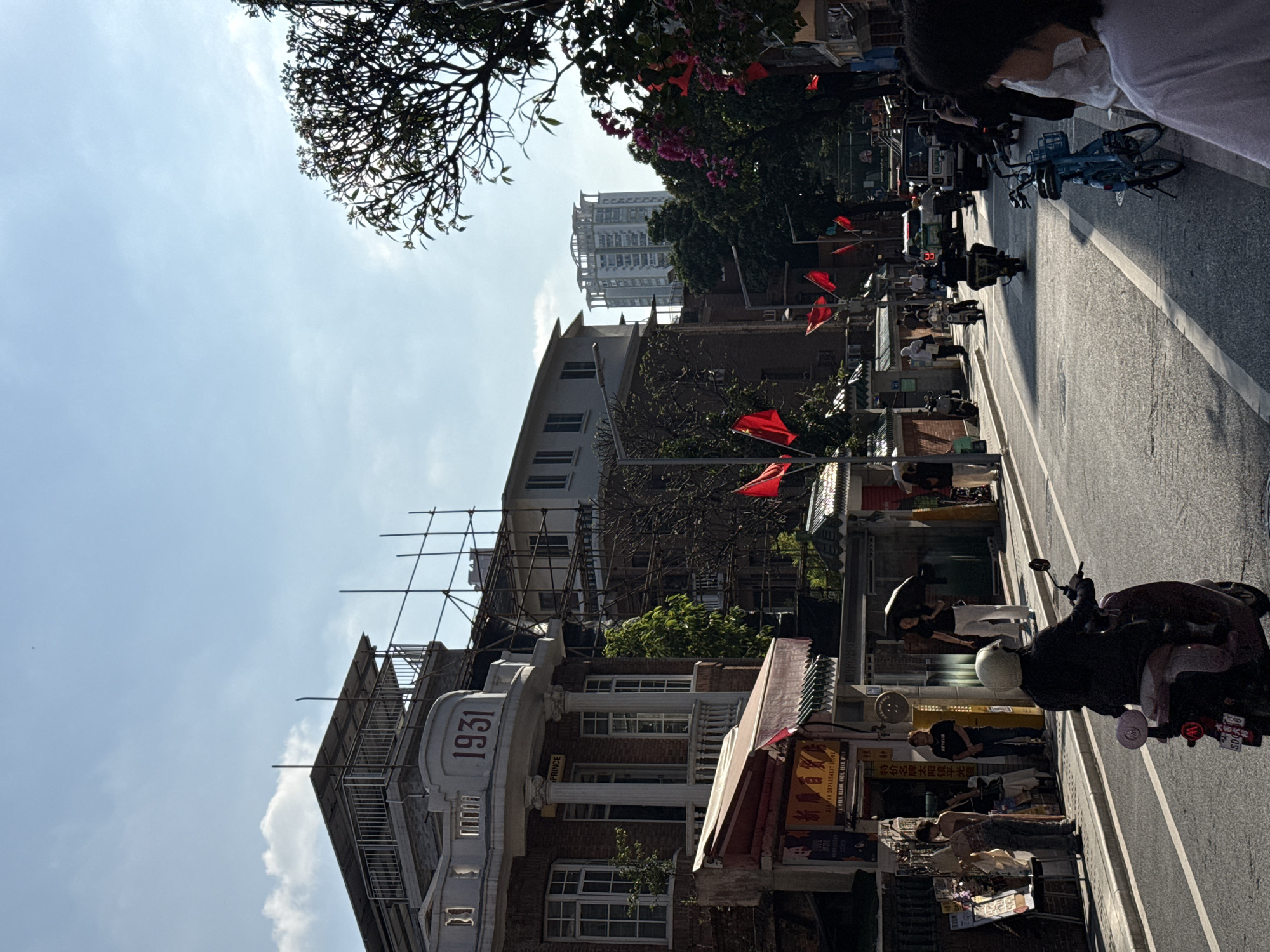 This image captures a vibrant street scene in an urban area, likely within Tianhe District, China. The perspective looks down a street bustling with daily activity under bright sunlight.
On the left side of the street, a mix of architectural styles is evident. A prominent building features classical elements like columns and balconies, with the year "1931" clearly visible on its facade. Below "1931", the word "PRINCE" is partially legible. Adjacent to it, another building is under renovation or construction, indicated by extensive scaffolding. Further in the background, a modern high-rise tower with a distinctive top stands tall, contrasting with the older structures. Trees with lush foliage and some with pink blossoms line parts of the street, and a large tree partially frames the top left of the clear, blue sky, which shows a few scattered clouds.
The street itself is paved, with a white line marking the edge of the road. Numerous red flags are displayed along the street, hanging from poles and buildings, adding a festive or commemorative element. The street is populated with people going about their day. Many are seen walking or standing near motorbikes and scooters, which are prevalent. A blue bicycle is parked further down the street. In the immediate foreground on the right, a person in a white shirt and a face mask is visible, observing the scene. Shopfronts with awnings line the street, displaying signs with Chinese characters. Visible text on signs includes characters for "Department Store" (商店) and "Tian Ji" (田记), among others.
The strong shadows and bright illumination suggest it is mid-morning or early afternoon on a clear, sunny day. The scene depicts a lively and typical urban environment, combining historical architecture with modern development and the daily routines of its inhabitants.