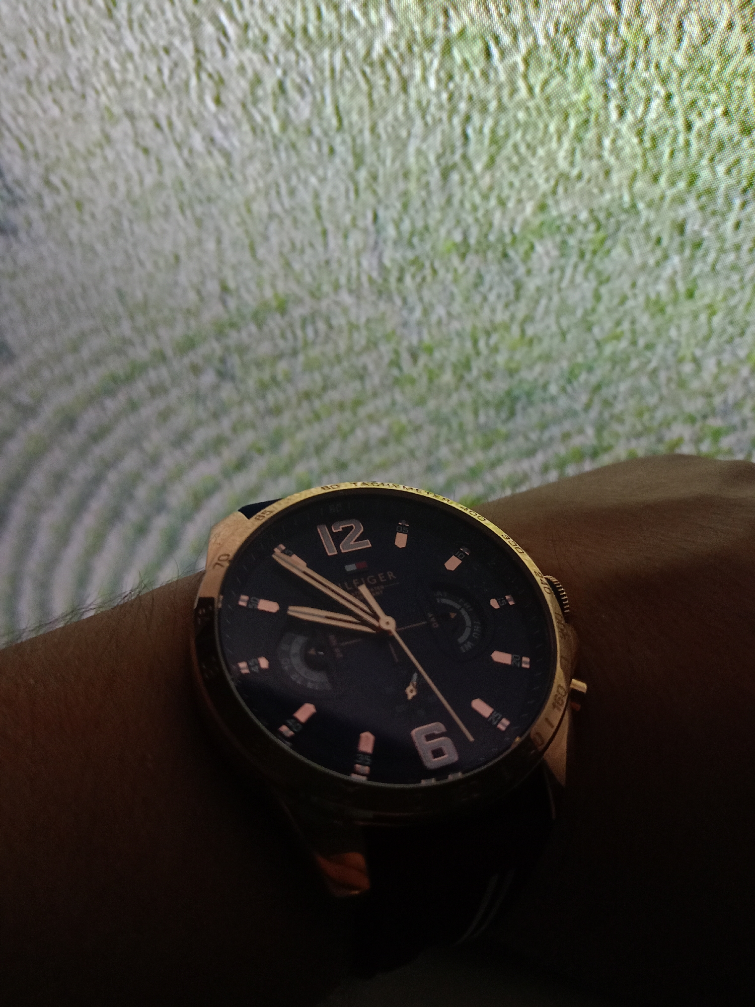 A close-up shot captures a rose gold Tommy Hilfiger watch on a wrist, against a blurred green background that appears to be grass or a similar textured surface. The watch face is dark blue with rose gold accents and hands. The hour markers are also rose gold, with Arabic numerals at the 12 and 6 o'clock positions. The watch is branded with "TOMMY HILFIGER" and features sub-dials, one of which displays the day of the week with abbreviations like "THU" and "FRI." The bezel has tachymeter markings. The lighting in the image is dim, creating shadows on the wrist and the lower portion of the watch. There are no identifiable features of Prostějov, Czechia in the image.