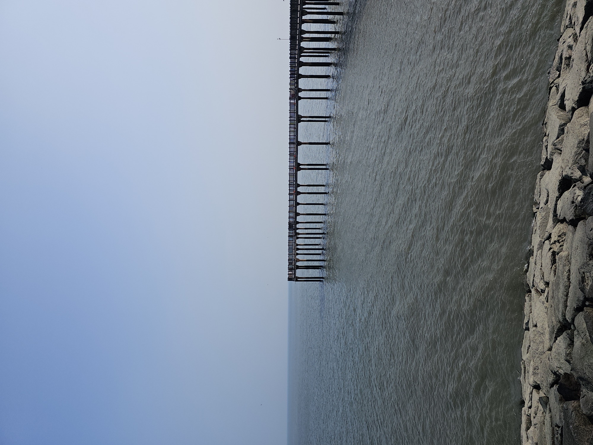 A long wooden pier extends out into the calm, grey water of the sea, supported by numerous dark, sturdy posts. The pier has railings along its length and appears to be empty of people. In the foreground, a rocky shore is visible, composed of large, grey stones. The sky above is a pale, clear blue, transitioning to a lighter hue near the horizon, where it meets the sea in a soft, diffused line. A few small birds are visible as specks in the sky and near the pier. The overall scene evokes a sense of tranquility and openness, with the pier serving as a man-made structure connecting the land to the vast expanse of the water. This is likely the Bangnyeongpo Pier, located in Hwaseong-si, South Korea, a coastal city known for its scenic waterfronts and piers.