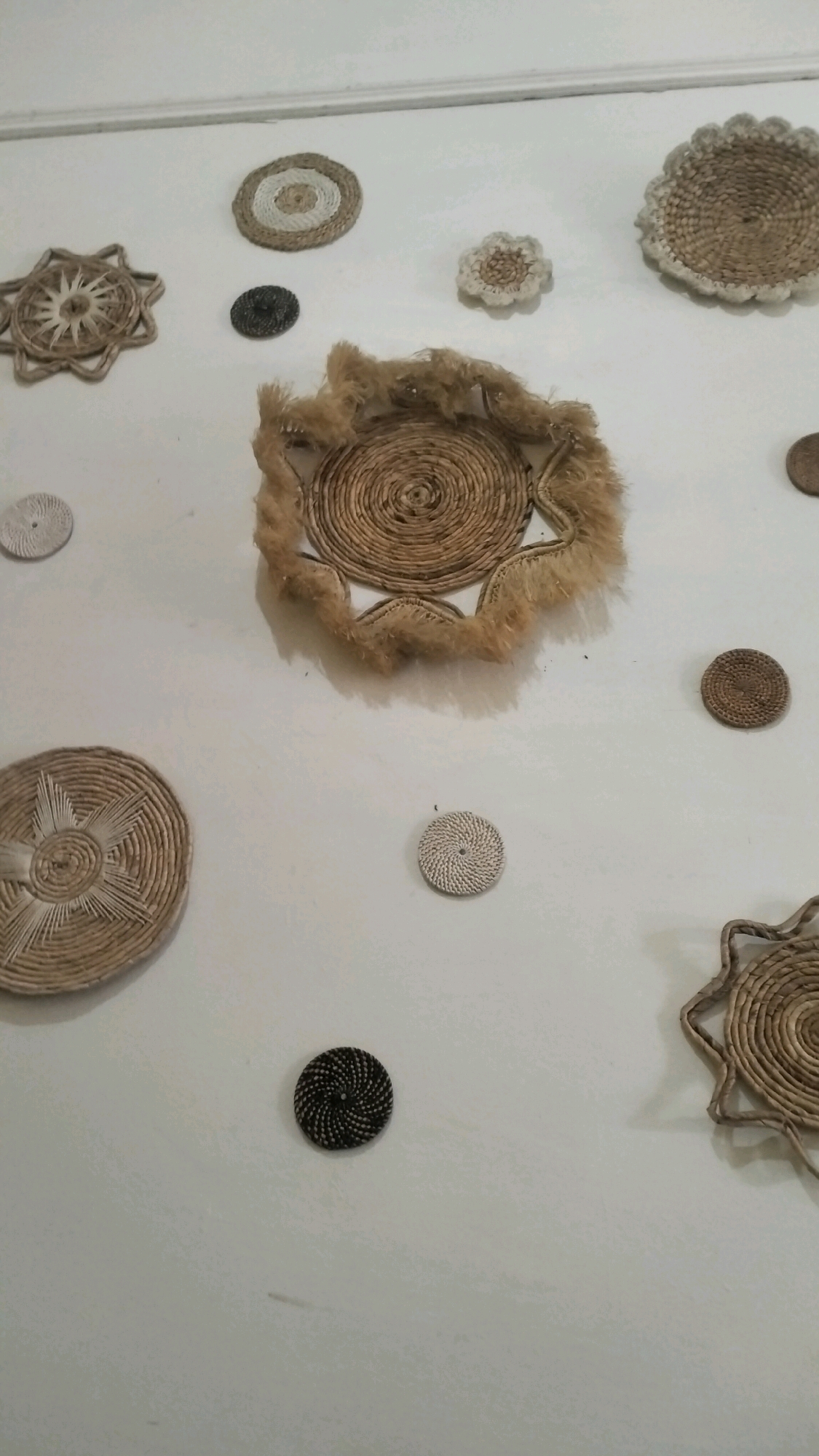 This image shows a collection of woven decorative items displayed on a white wall, likely in a home or shop in Lhokseumawe, Indonesia. The items are made from natural fibers such as straw or reeds, and vary in size and design. They include circular mats with intricate patterns, some with star-like or floral shapes, and one larger, shallow bowl-like item with a fringe of fibers. The lighting appears to be natural or soft artificial light, casting subtle shadows on the wall. There are no people visible in the image. The setting is simple and uncluttered, focusing attention on the handcrafted decor.
