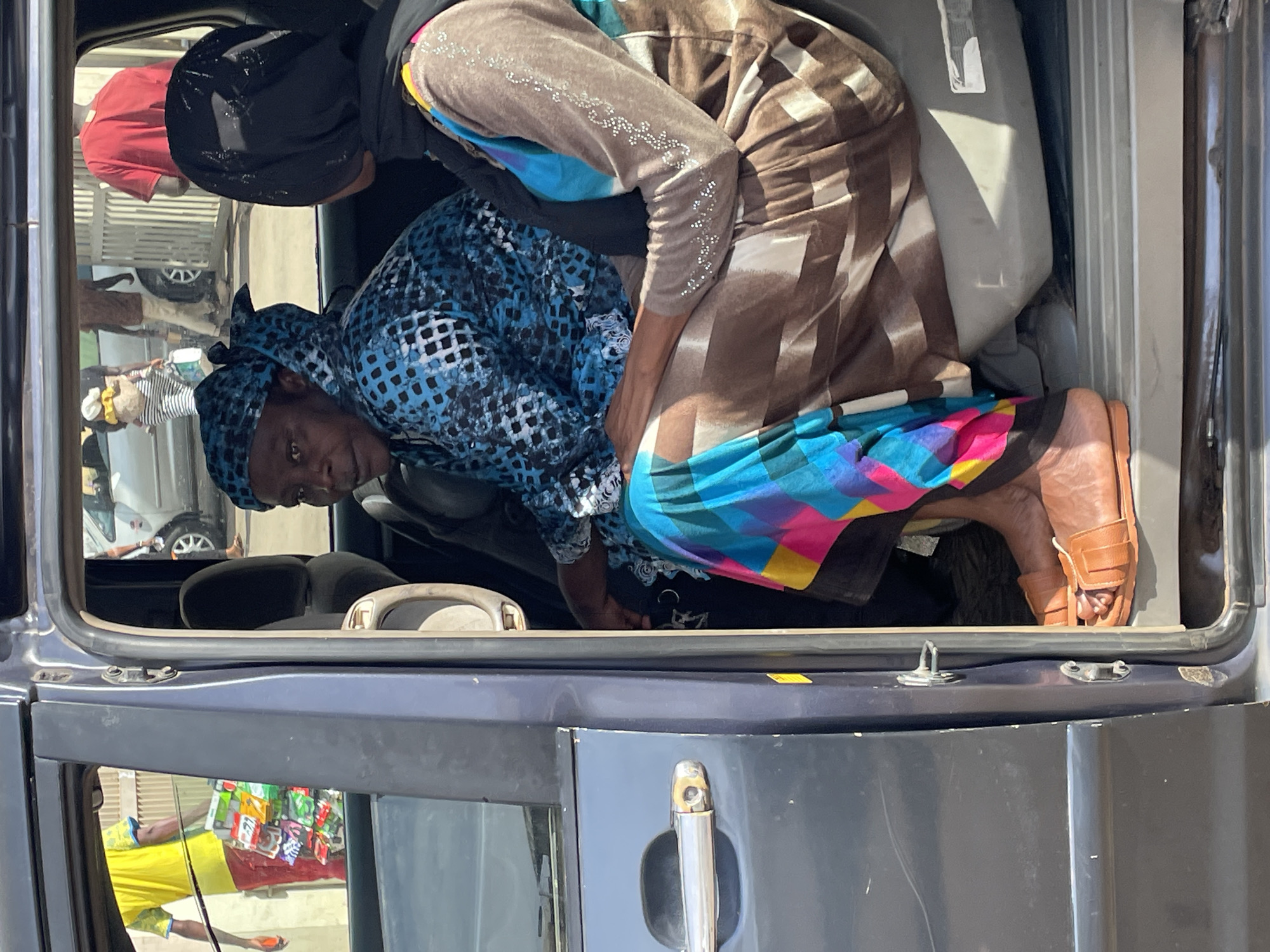 In Bukuru, Nigeria, two African women are seen inside the open back of a dark blue van. One woman, wearing a blue patterned dress and headscarf, is crouched down, looking into the van's interior. The other woman, wearing a brown and white striped outfit with a black head covering, is bent over, seemingly assisting the first woman. Both women are barefoot, with one woman wearing brown sandals. Inside the van, a white object is visible, possibly luggage or supplies. The exterior of the van is visible, with a door handle and a window reflecting a vendor's stall. The background includes a glimpse of a street scene with other vehicles and people.