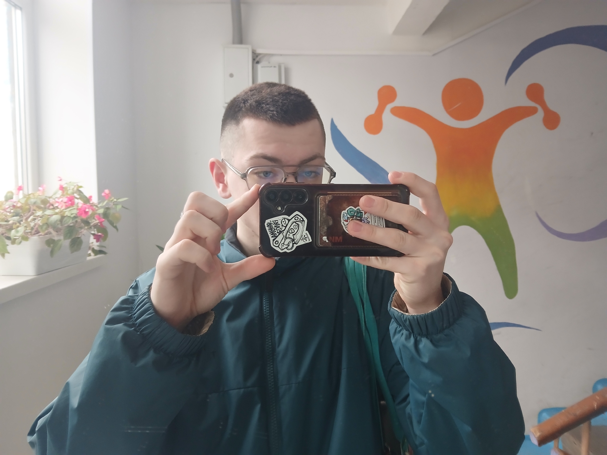 A young man wearing glasses and a dark green jacket is taking a selfie with his phone. The phone has stickers on the back. He is indoors, standing in front of a white wall with a colorful abstract mural of a person with outstretched arms under a blue crescent shape. To the left is a window with a flower pot containing pink flowers. The location is in the city of Chaikovsky, Russia.
