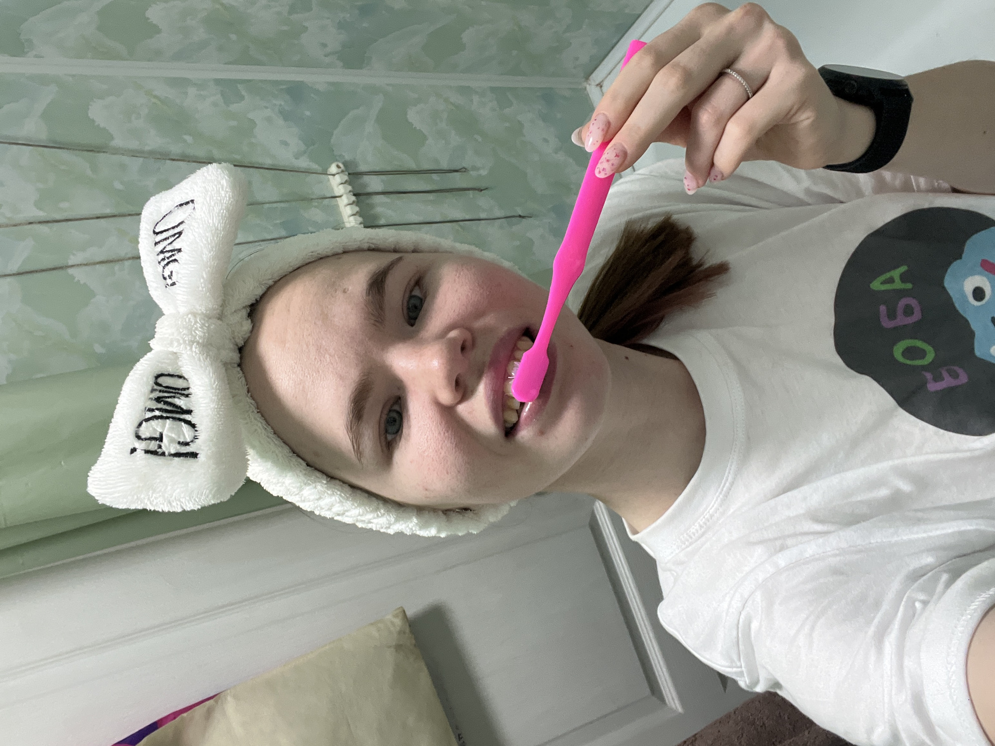 A young woman is brushing her teeth with a pink toothbrush. She is wearing a white headband with "OMG!" written on it and a white t-shirt with a cartoon graphic. She has a black smartwatch on her left wrist and a ring on her finger. The background appears to be a bathroom with a light green wall and a white door. The location may be in Chaikovsky, Russia, as specified in the prompt.