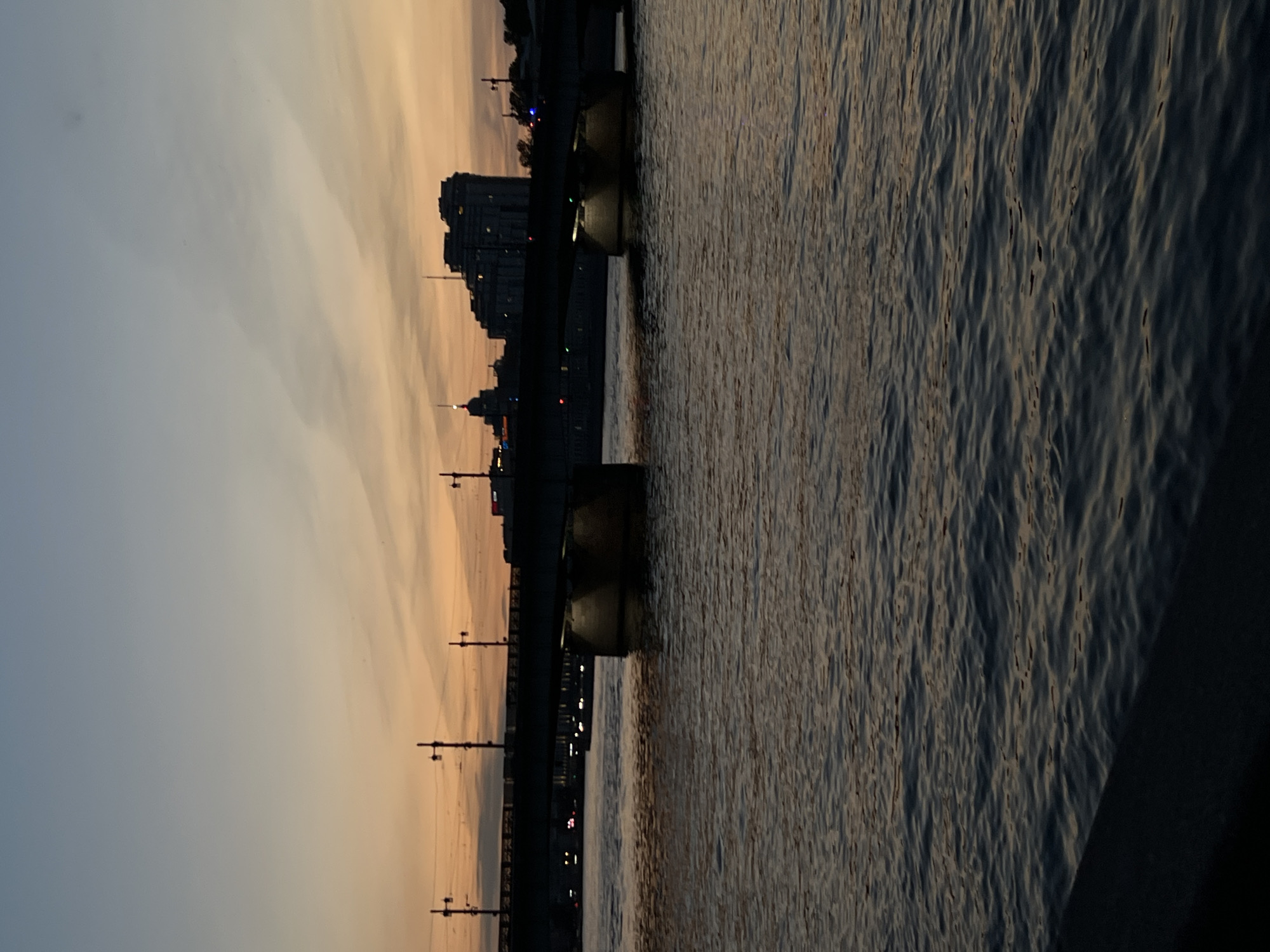 The image depicts a city skyline at dusk. A bridge spans across a body of water, with the silhouettes of buildings visible behind it. Several streetlights stand along the bridge. The water is dark and rippled, reflecting the dim light of the sky. The sky is a gradient of orange and blue, with wispy clouds. The scene suggests an urban environment at the transition from day to night.