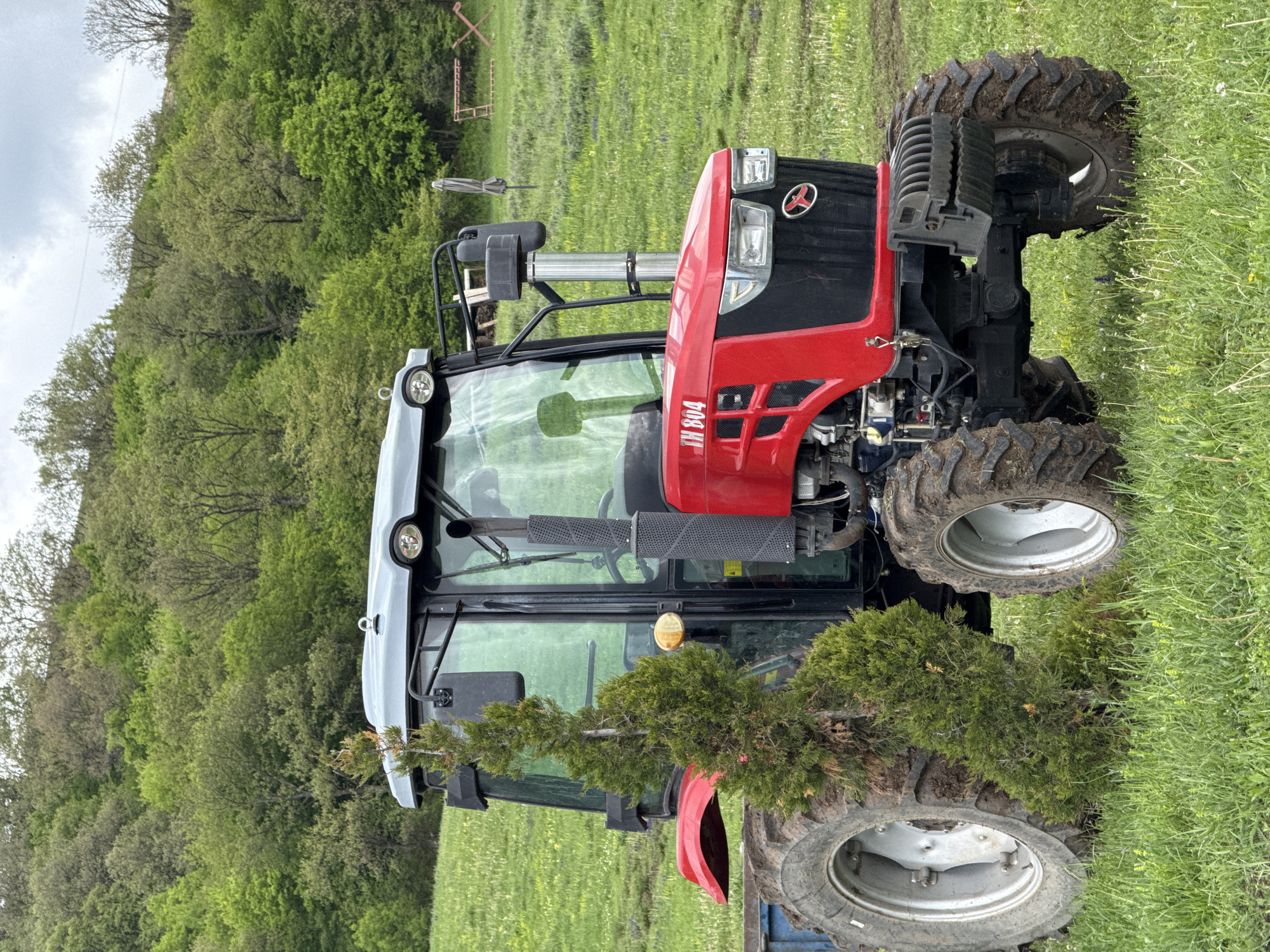 A red and black tractor is parked in a grassy field. The tractor has large, mud-covered tires and a white cab. Some small evergreen trees are on the tractor. In the background, there's a lush green hillside and some agricultural equipment. The location is Dsegh, Armenia.