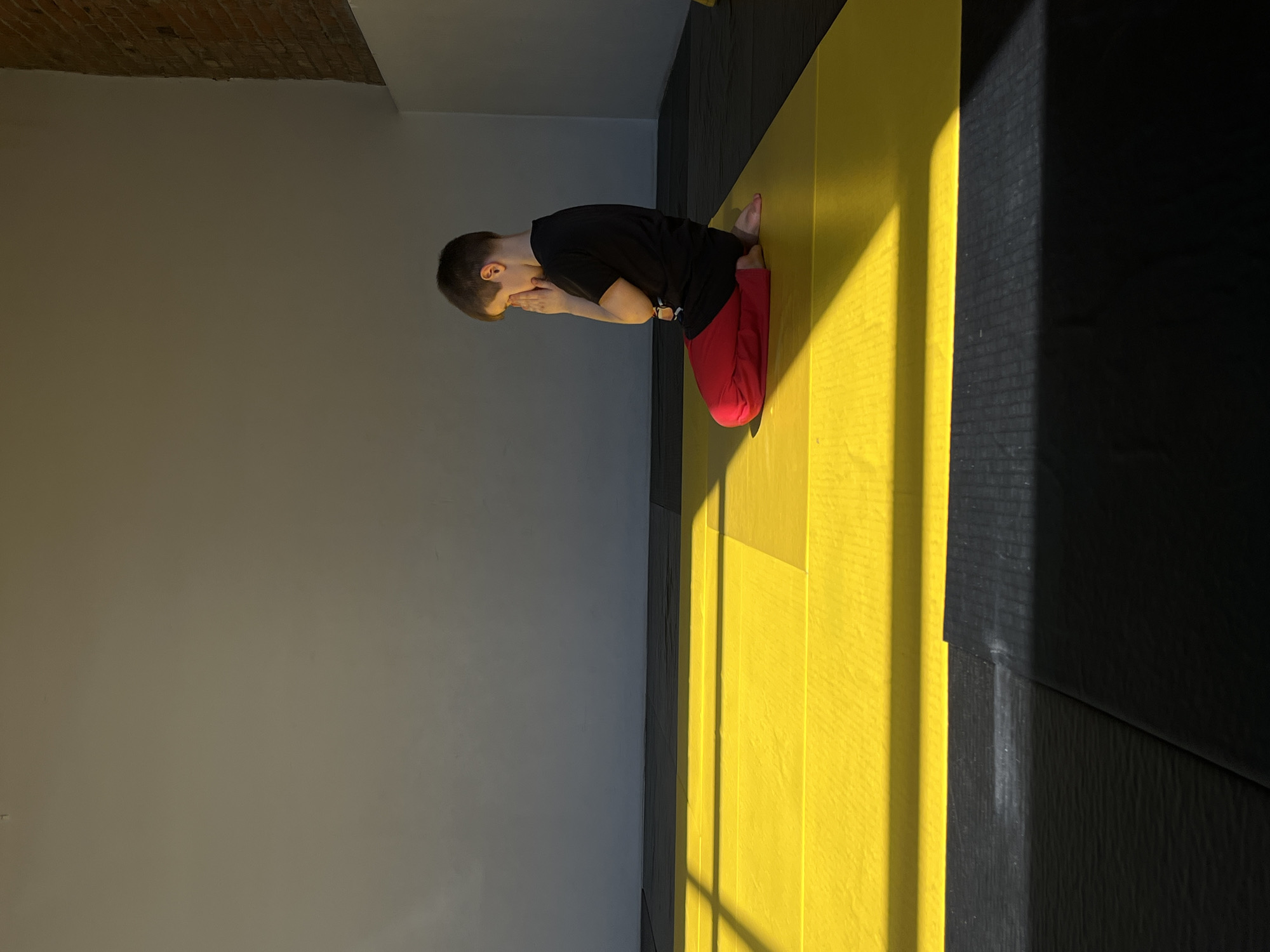 The image shows a young person kneeling on a yellow and black floor in a room with grey walls and a brick ceiling. The person is wearing a black shirt and red pants, and their head is bowed, with their hands near their face. The lighting is strong, with a distinct shadow pattern on the yellow floor. There is no indication of the location being in Krasnoyarsk, Russia, or any specific city.