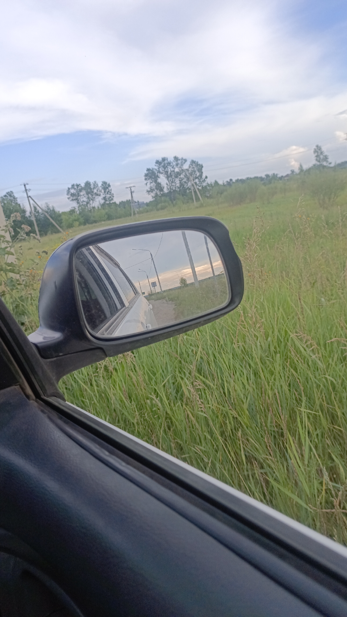 The image shows a view from the passenger side of a car looking out at a rural landscape. In the foreground, the dark interior of the car's door is visible. To the right, tall green grass lines the side of the road. In the center of the image, a side-view mirror reflects the scene behind the car. The reflection shows a light-colored car, a dirt road, power lines with poles, and a treeline under a cloudy sky with patches of sunlight. In the background, beyond the grass and trees, there are more power lines and poles. The overall impression is that of a journey through a rural area on a cloudy day. The reference to Angarsk, Russia, suggests the geographical context, but there are no specific landmarks or elements in the image that definitively place it within the city of Angarsk.