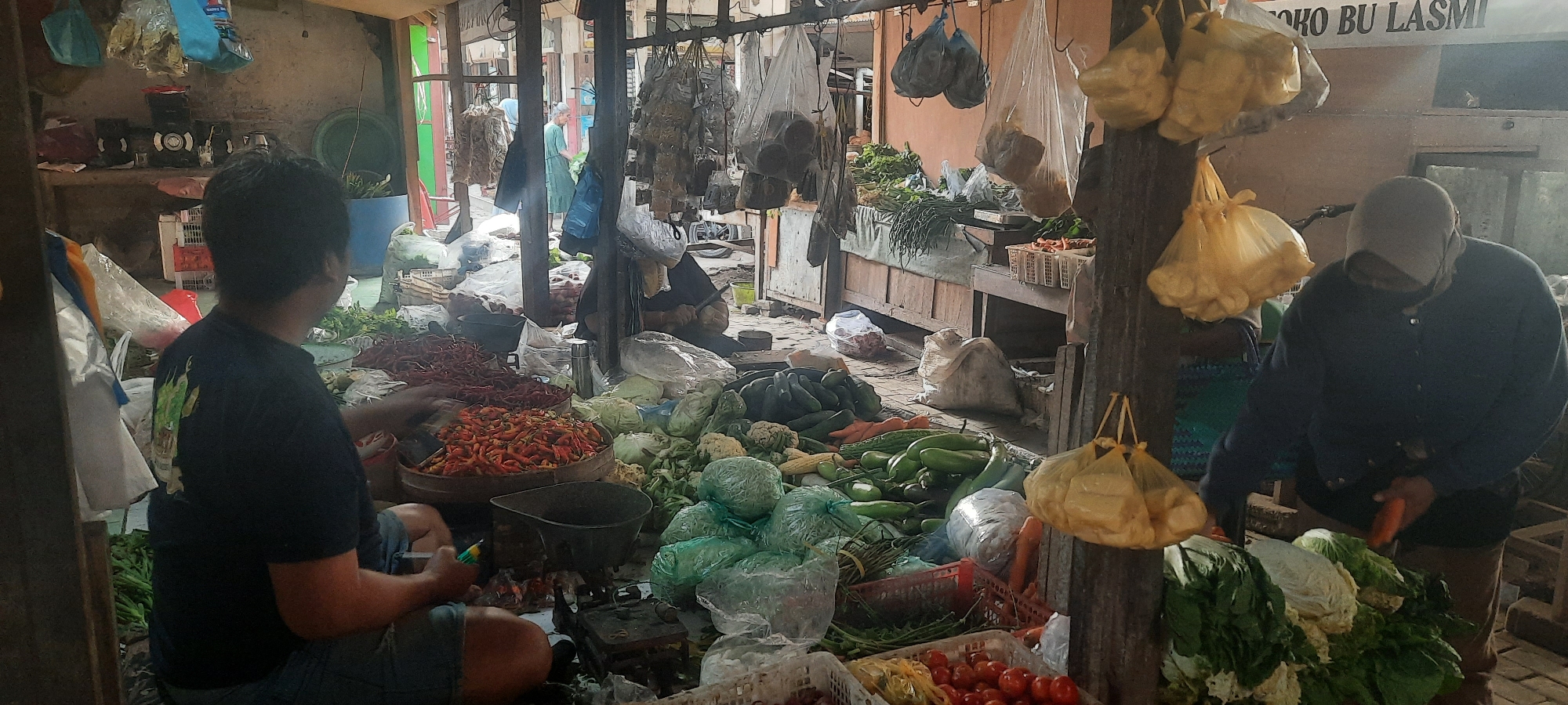 This image depicts an outdoor market scene in Selopuro, Indonesia. In the foreground, a person wearing a black t-shirt and blue shorts is seated and appears to be sorting through a pile of red chili peppers. To their right, a second person, wearing a dark blue jacket and a light-colored head covering, is seated and is seen holding and cutting an orange vegetable, likely a carrot. The market is filled with a variety of fresh produce, including more chili peppers, cabbages, cauliflower, cucumbers, and tomatoes. Several plastic bags containing produce are hanging from overhead structures. In the background, there are wooden stalls and structures that are typical of a market environment. The overall scene suggests a busy marketplace with vendors and their goods on display.