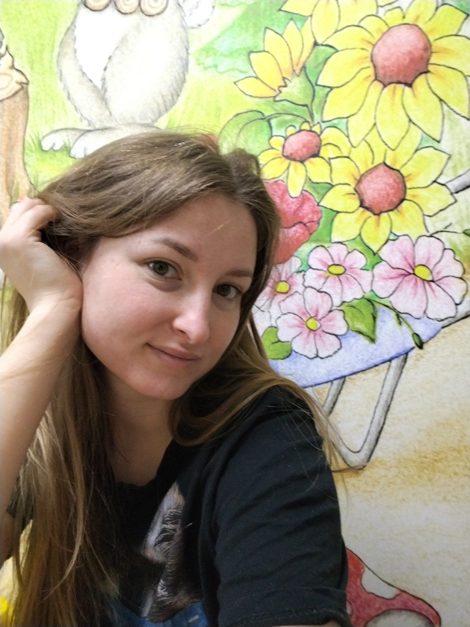 The image captures a young adult female in the city of Chaikovsky, Russia. She possesses a light complexion, long light-brown hair, and green eyes, with a slight smile directed at the viewer. Her left hand is positioned with fingers resting within her hair, near her left ear. She is dressed in a black short-sleeved t-shirt with a faint graphic, possibly an animal design, on the chest. The background is a brightly colored wall mural or wallpaper. Visible elements of this mural include a partial grey animal figure with circular features in the upper left, a large cluster of yellow sunflowers with red centers, and pink flowers with yellow centers, some arranged in a blue container, on the right side. A segment of a red and white mushroom is also visible in the lower right background. The lighting appears even and consistent with an indoor setting.