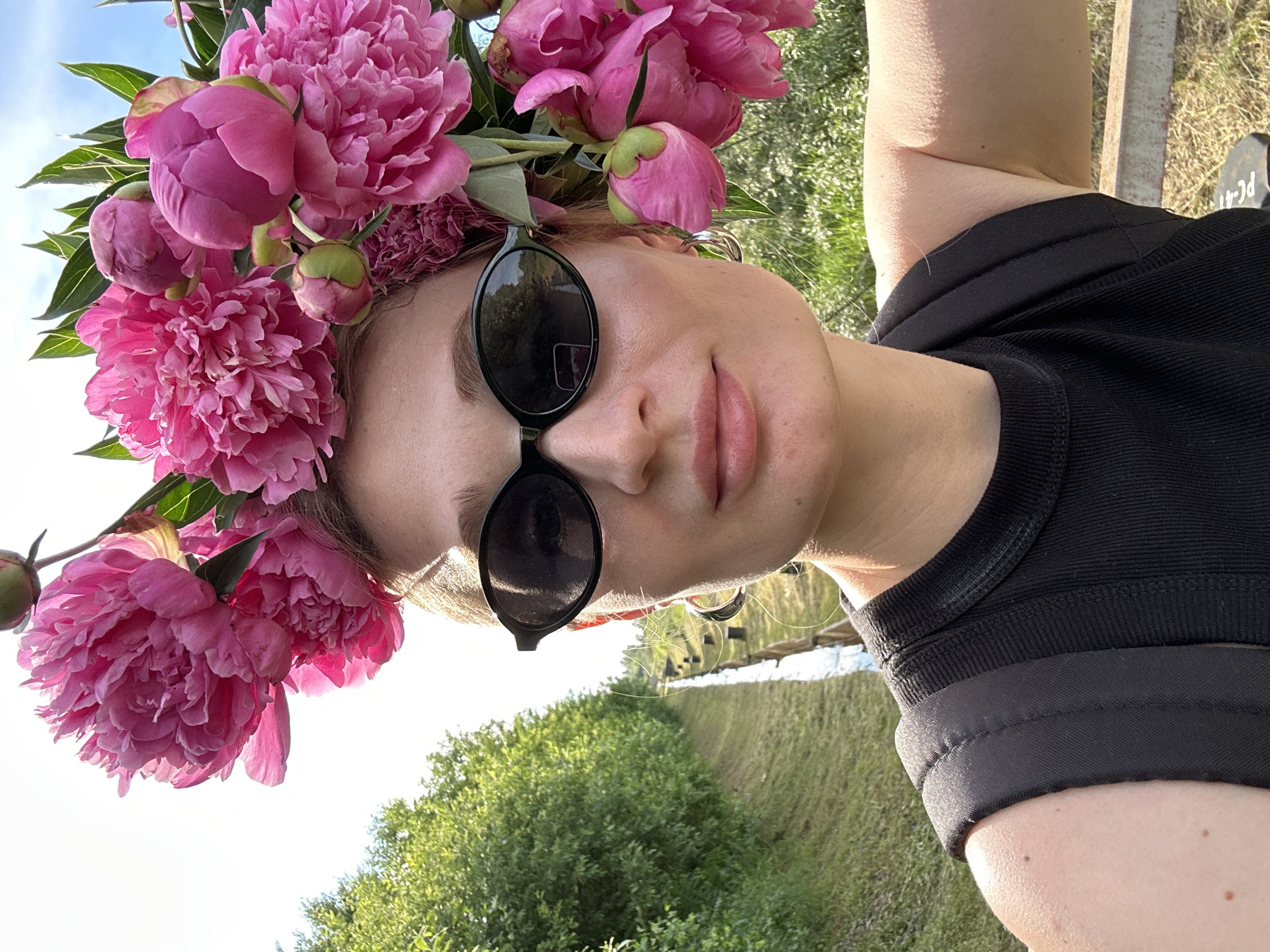 The media file is a close-up, vertically oriented selfie of a person wearing a crown of pink peonies. The subject, who appears to be a young woman, is wearing dark sunglasses and a black top. The background is blurred, but hints at an outdoor setting with greenery and a body of water, possibly a canal or river, suggesting a sunny day. The lighting indicates it is likely daytime, with the sun casting shadows. The overall mood seems relaxed and joyful, with the floral crown adding a whimsical and festive touch. The image is taken at a low angle, with the subject's face tilted upwards towards the camera, and the peonies dominating the upper portion of the frame. The subject's skin tone is fair, and she has subtle makeup on. There are no discernible text elements within the image. The context provided, Chaikovsky, Russia, suggests the natural environment shown is consistent with this location, likely during the warmer months when peonies would be in bloom.
