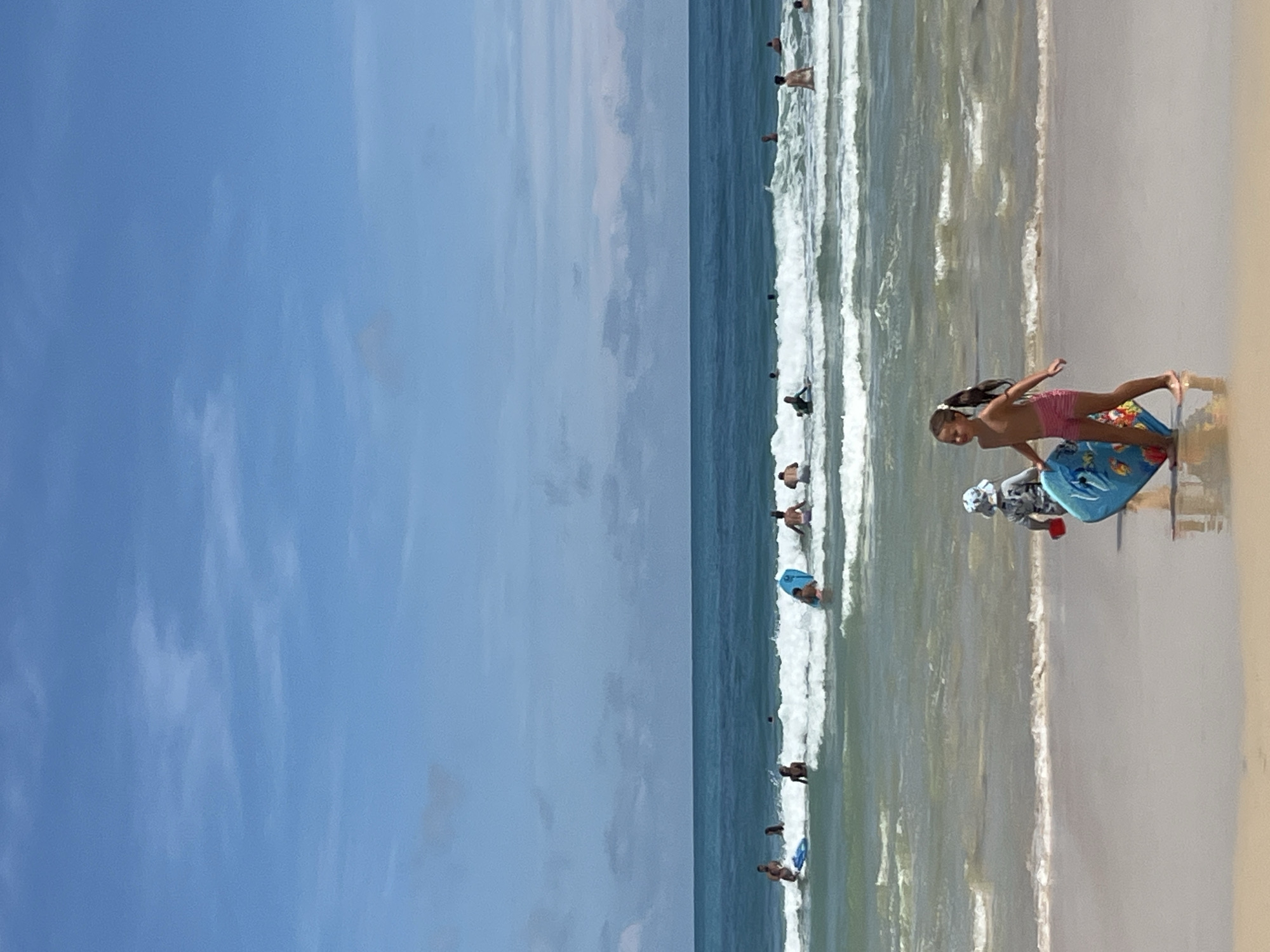 A vertical, medium shot depicts a bright, sunny day at a beach in Rawai, Thailand, with the Andaman Sea and a clear blue sky. In the foreground, two children are on the wet sand near the water's edge. A girl, wearing pink shorts, is walking away from the camera to the right, carrying a blue boogie board. Beside her and slightly behind, a younger child in a sun hat and patterned long-sleeved shirt is also holding a blue boogie board and a red toy shovel. The water is a vibrant turquoise with white crests on the waves. Further out in the sea, several people are visible, some swimming or wading, and others are on surfboards or boogie boards in the shallow waves. The horizon is a sharp line separating the sea from the sky. The sky is mostly clear with scattered white clouds. The overall scene conveys a typical beach day with recreational activities.