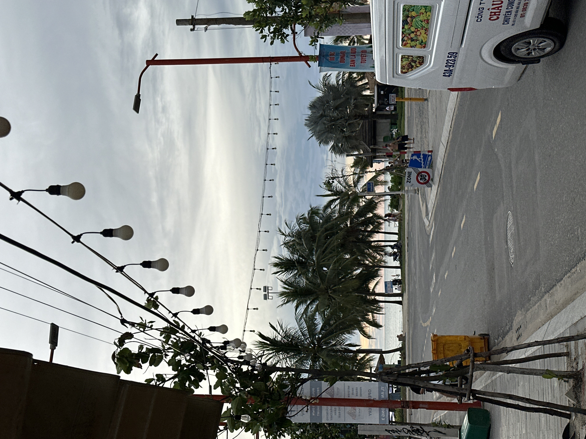 The image depicts a street scene in Đà Nẵng, Vietnam, likely near the beach given the presence of palm trees and the ocean visible in the background. The sky is overcast with a gradient of gray, suggesting either early morning or late afternoon. Overhead, a string of decorative light bulbs is strung across wires, with a street lamp on a red pole also visible.
On the left side of the street, a building's facade is partially visible, with a small tree supported by stakes in front of it. A bright yellow trash bin and a green rectangular bin are also present on the sidewalk.
The street itself is asphalt with visible lane markings. A white van with the license plate "43A-923.59" and Vietnamese text is parked on the right side of the road. Signs are posted along the sidewalk, including a "ZONE" sign with a pedestrian symbol and a speed limit indication.
In the middle ground, several people are walking and standing, some near a cluster of palm trees and a low-lying structure. The ocean and beach are visible beyond the trees, with a few individuals and motorcycles present near the water's edge. The overall atmosphere is that of a coastal urban environment.