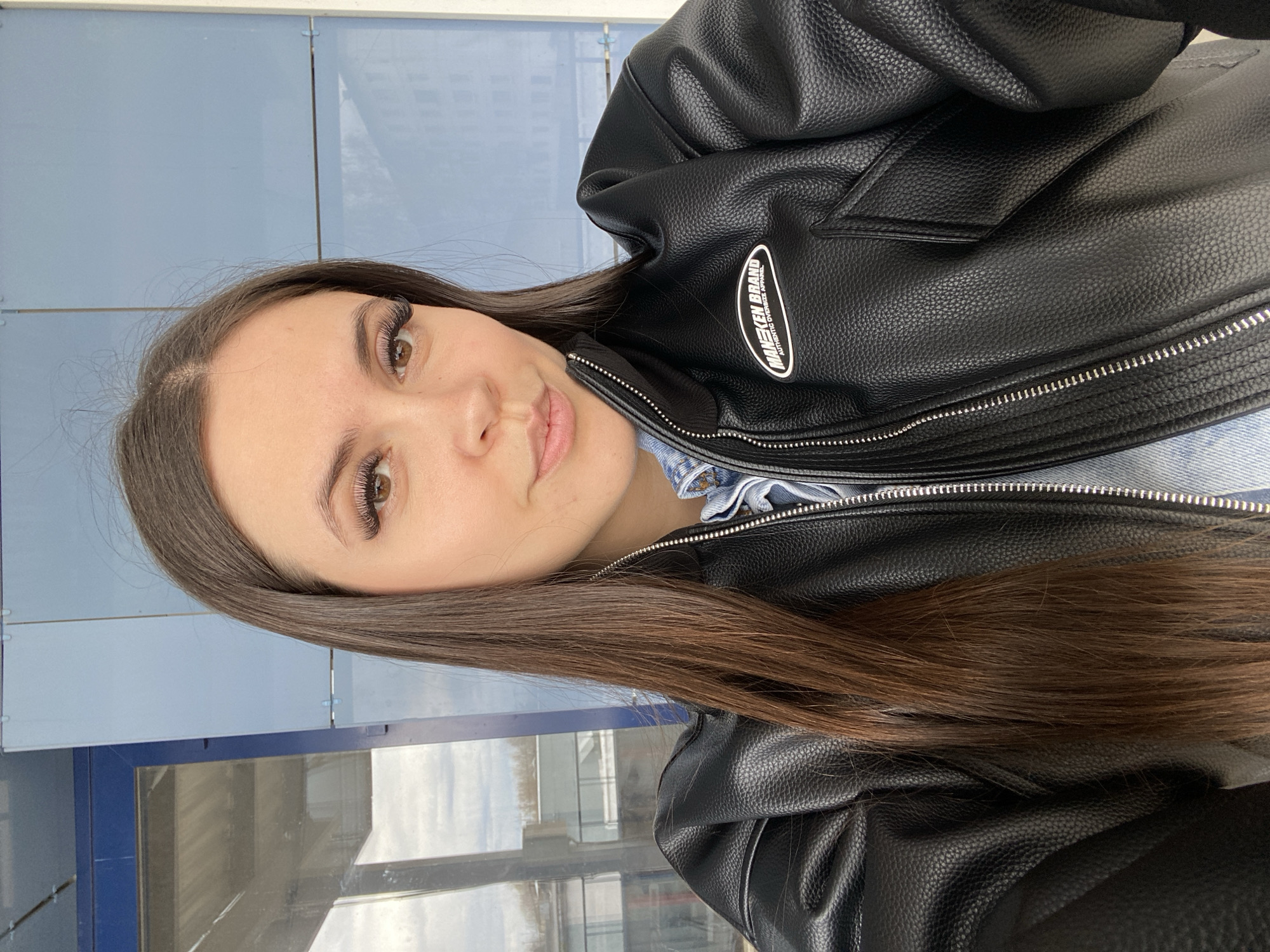 The image displays a head-and-shoulders portrait of a young woman, likely a selfie, taken from a slightly elevated angle. She has long, straight, dark brown hair that flows down the left side of the frame and over her shoulders. Her brown eyes are accentuated by prominent, dark eyelashes, and she has a subtle, closed-mouth smile on her naturally colored lips.
She is wearing a black, textured faux leather jacket with a silver zipper running down the front. The jacket features a white oval patch on her right chest area (viewer's left) with black text. The text reads "MAI KIKEN BRAND" with "AUTHENTIC OVERSIZED APPAREL" written below it in smaller letters. Underneath the jacket, a light-colored, possibly denim, shirt or top is visible around her neck and chest area, showing some gathering or ruffling.
The background primarily consists of light blue-grey panels or walls, indicative of an indoor or semi-enclosed public space. Below these panels, large glass windows or reflective surfaces are visible, reflecting a bright, possibly overcast sky and vague outlines of distant structures or buildings. The lighting suggests it is daytime. The woman appears calm and content, looking directly at the camera. The setting points to a contemporary indoor environment, possibly a station or large building in Chaikovsky, Russia.