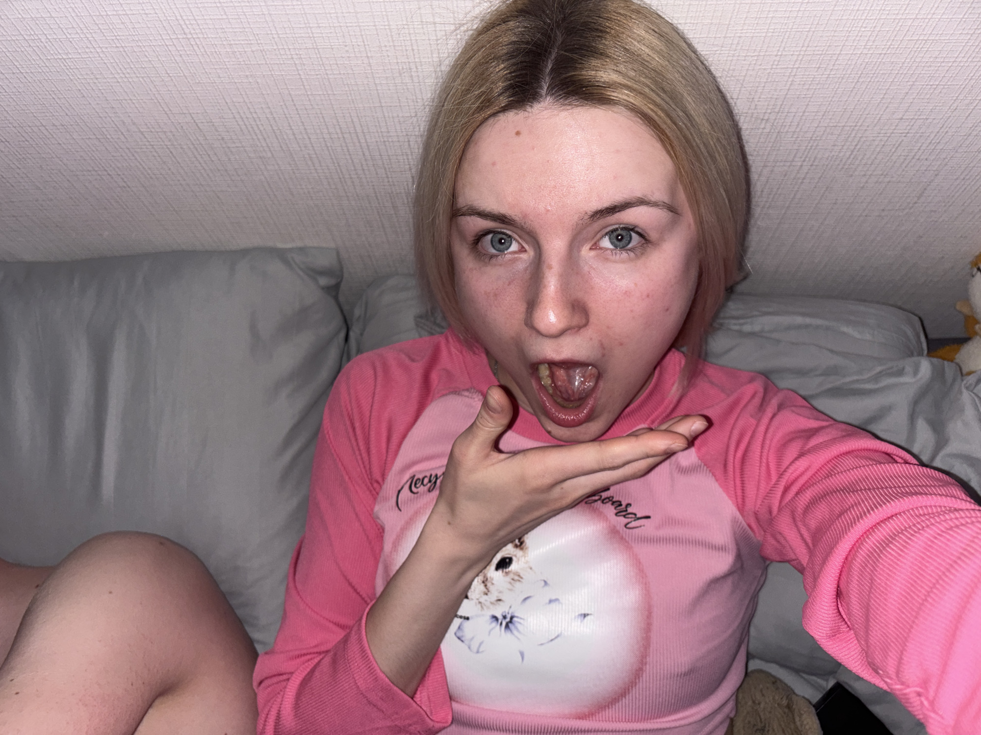 A young woman with blonde hair and blue eyes is featured in a close-up, informal selfie taken indoors. She is positioned against a gray pillow and bedding, with a textured white ceiling visible above her.
The woman wears a long-sleeved pink raglan-style shirt. The shirt has a circular white graphic on the front depicting a fluffy cat, with faint text partially visible around it, including "Recy" and "My board". Her mouth is wide open, tongue extended, and her eyes are wide, conveying a playful or surprised expression. Her left hand is held open, palm up, below her chin. She has natural skin with some redness and minor blemishes on her face, and her blonde hair shows darker roots.
In the upper right background, a small, light-colored plush toy is partially visible. The indoor setting, likely illuminated by artificial light, suggests a casual environment such as a bedroom or living area. The image captures a candid moment in Ligolambi, Russia.