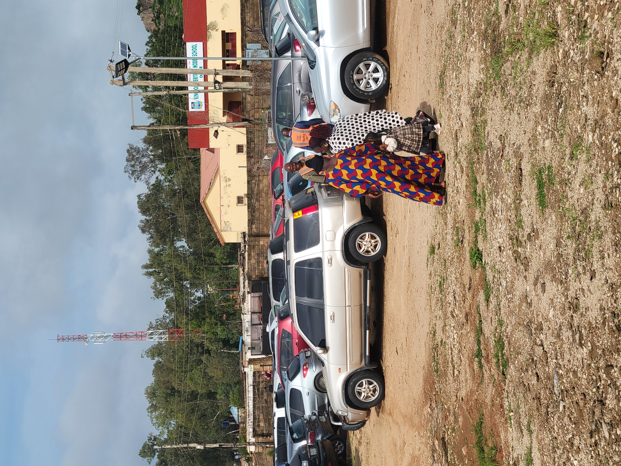 The image depicts an outdoor scene in the city of Gura Top, Nigeria, featuring a parking lot filled with various cars. A prominent cell tower stands in the background, suggesting a well-connected area. A school building, identified by a sign that reads "KINGDOM HERITAGE MODE SCHOOL," is also visible, indicating a place of education. Several individuals are present, including a man wearing an orange vest with the inscription "ESC TRAFFIC CONTROL UNIT." He appears to be interacting with a group of women and a child dressed in colorful traditional attire. The ground is predominantly dirt and gravel, with some sparse vegetation, consistent with an outdoor setting in a warm climate. The overall scene suggests a busy day at the school or a local event.