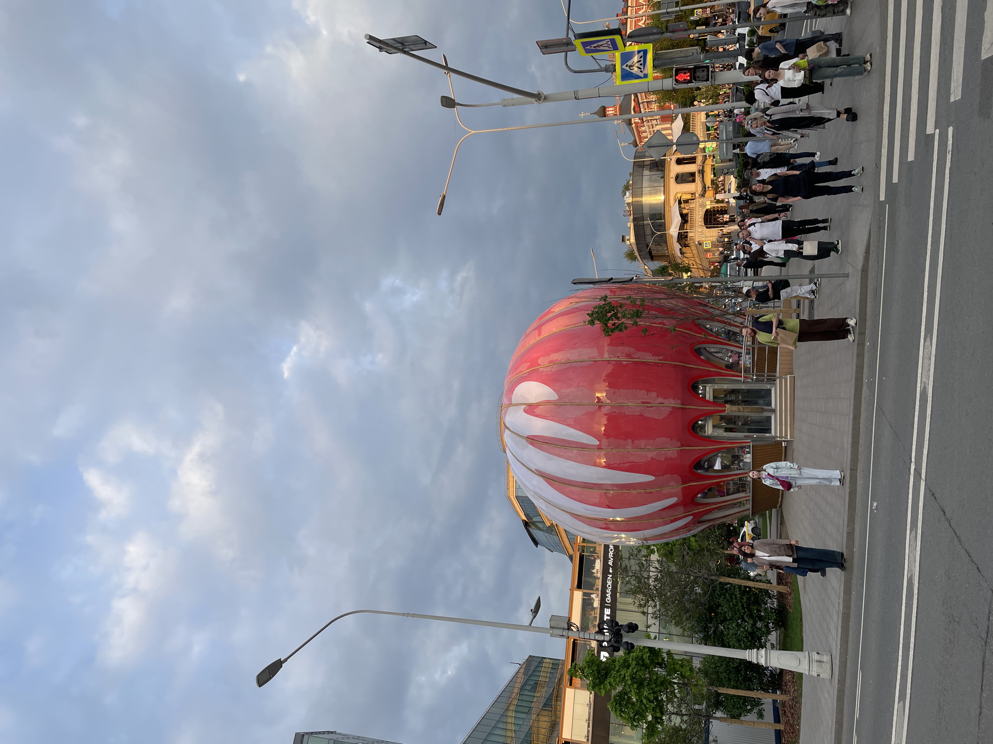 The image depicts a street scene in Moscow, Russia. A large, spherical red building with white swirls is prominently featured. The building has a wooden structure and appears to be a commercial establishment, possibly a cafe or shop. Pedestrians are present on the sidewalk, including a group of people crossing a crosswalk. Streetlights and traffic signals are visible. The sky is overcast with grey clouds. Buildings and greenery flank the street. The facade of a building in the background displays the text "GARDEN OF AVROP".