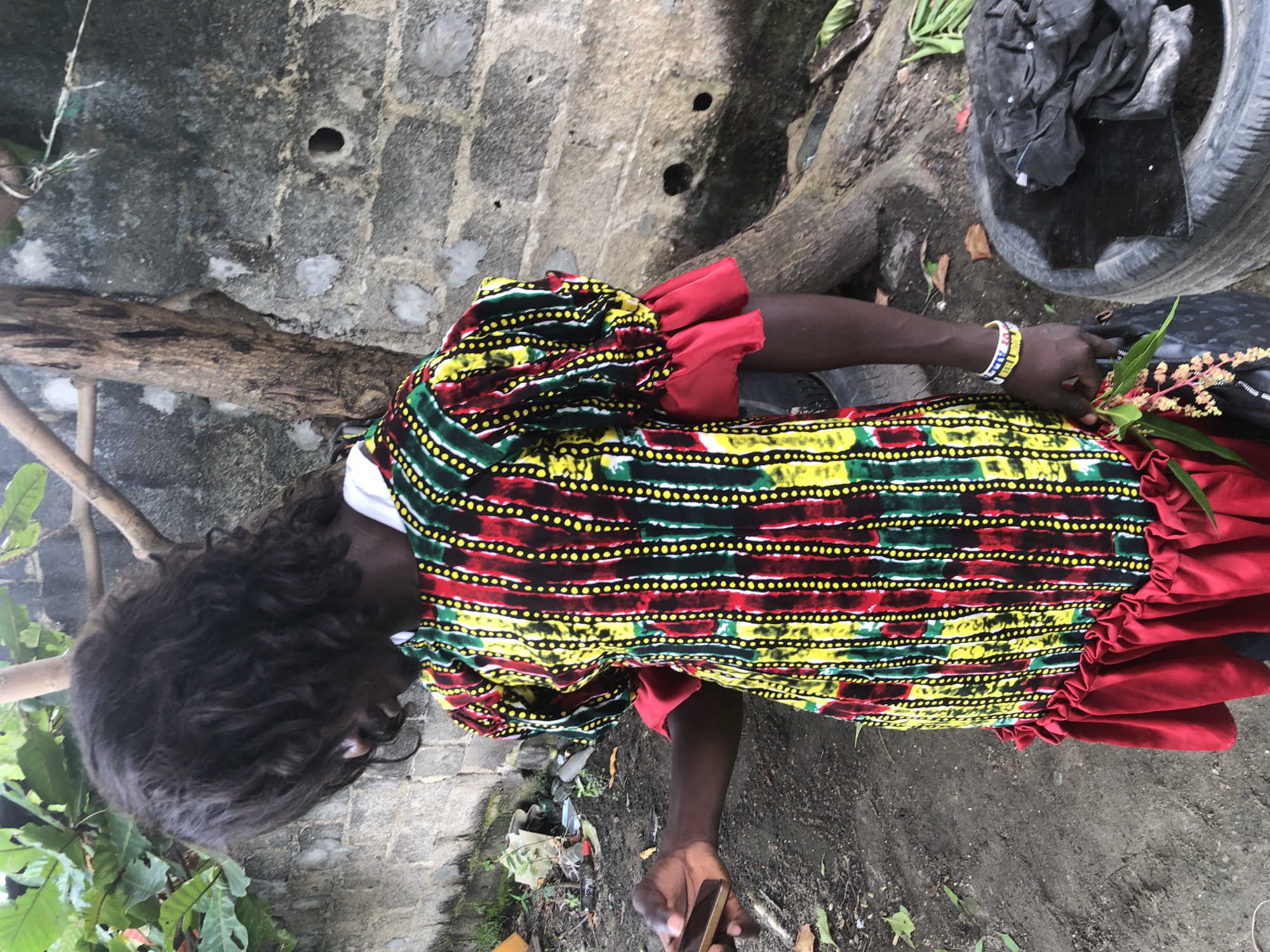 A person stands in front of a rough, grey brick wall with green foliage and tree branches above. The person is wearing a vibrantly patterned dress with vertical stripes of yellow, red, and green, adorned with small yellow dots. The dress has ruffled sleeves and a ruffled hemline. The person's left hand holds a smartphone, while their right arm displays a beaded bracelet. In the background, to the right of the person, a large tree trunk is visible, and at the bottom right, a used tire filled with dark, crumpled material. The ground appears to be dirt and debris. The scene is likely an outdoor, urban setting within Ado, Nigeria, given the context provided.