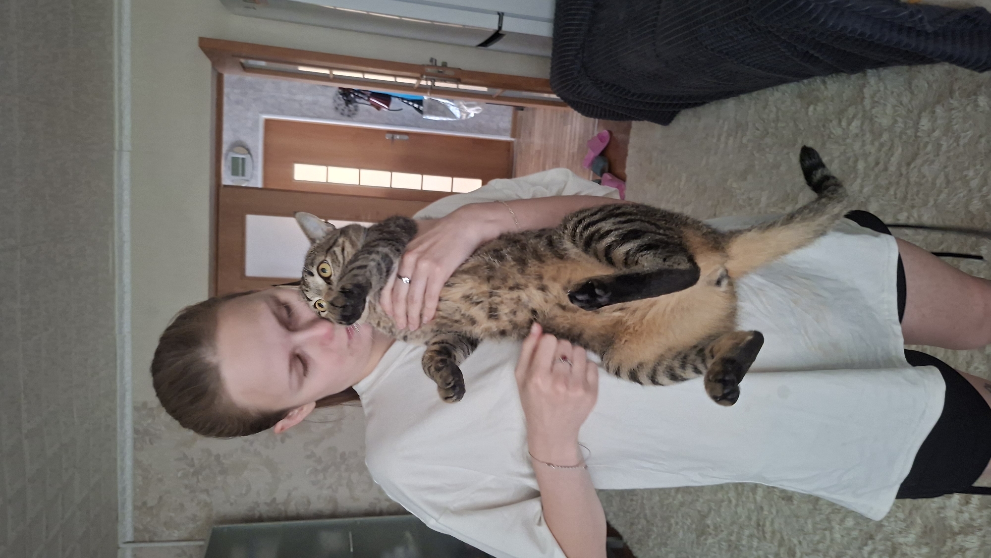 A young woman with brown hair pulled back in a ponytail is holding a tabby cat in her arms. The cat is facing the camera, with its paws up near its face. The woman is wearing a white t-shirt and black shorts. The scene appears to be indoors, in what looks like a living room. A doorway with a glass panel is visible in the background, as is a sofa covered with a dark blanket. The floor is covered with a light-colored rug. The overall impression is a casual, domestic setting. There is no information to suggest the location is in Myadzyel, Belarus.