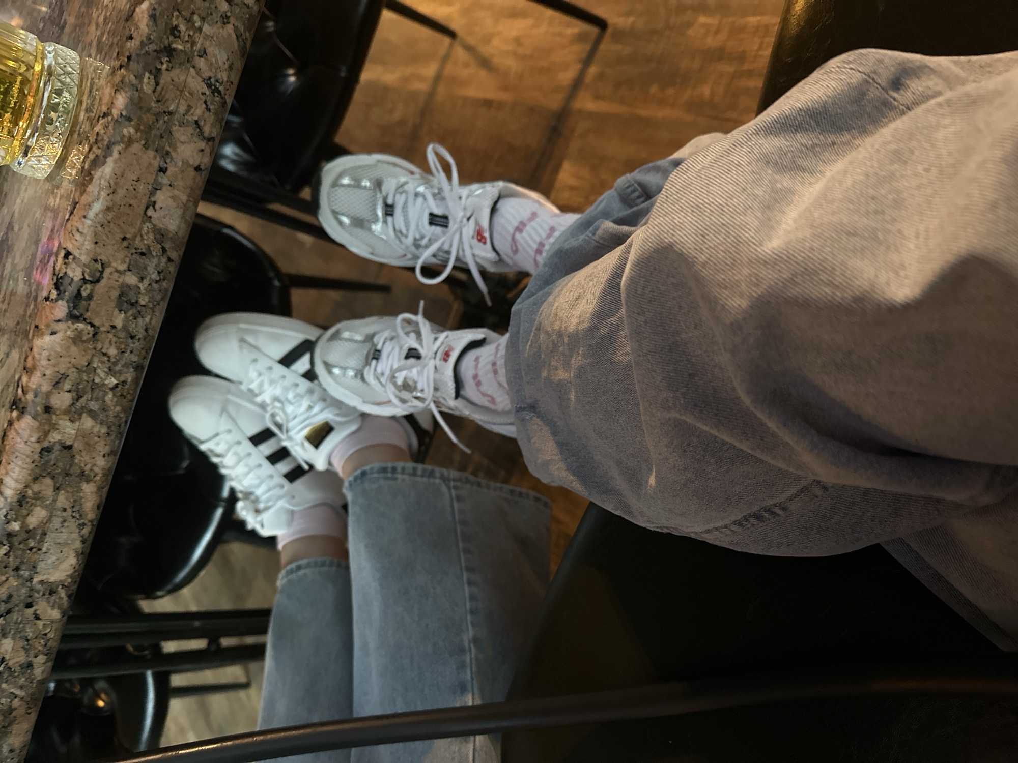 The image shows a person sitting at a bar with a drink. The person is wearing light-wash jeans and two different pairs of white sneakers: one pair of New Balance sneakers and one pair of Adidas sneakers with black stripes. The bar has a dark, textured surface and is set on black bar stools. The location is not specified as being Votkinsk, Russia.