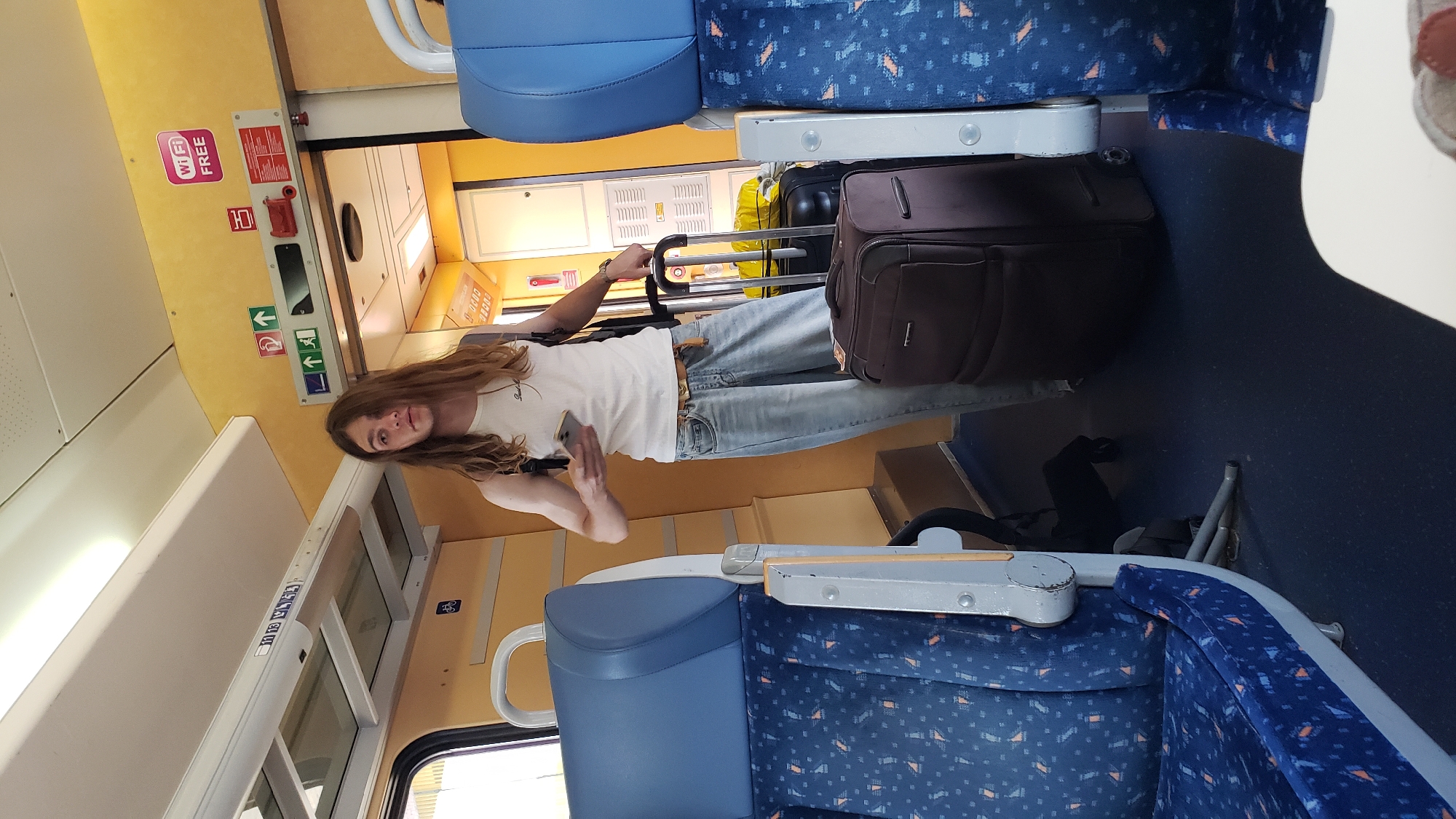A young person with long, light brown hair stands in the aisle of a train carriage, holding a smartphone in their right hand. They are wearing a white tank top and light-wash, loose-fitting jeans. A brown suitcase is positioned to their right, and another dark suitcase is partially visible behind it. The train interior features blue seats with an orange and blue patterned upholstery, overhead luggage racks, and yellow walls. Signage on the wall indicates "Wi-Fi FREE" and directional arrows. The scene appears to be within a train traveling to or from Žilina, Slovakia, given the presence of the "Wi-Fi FREE" sign, which is common in Slovakian public transport.