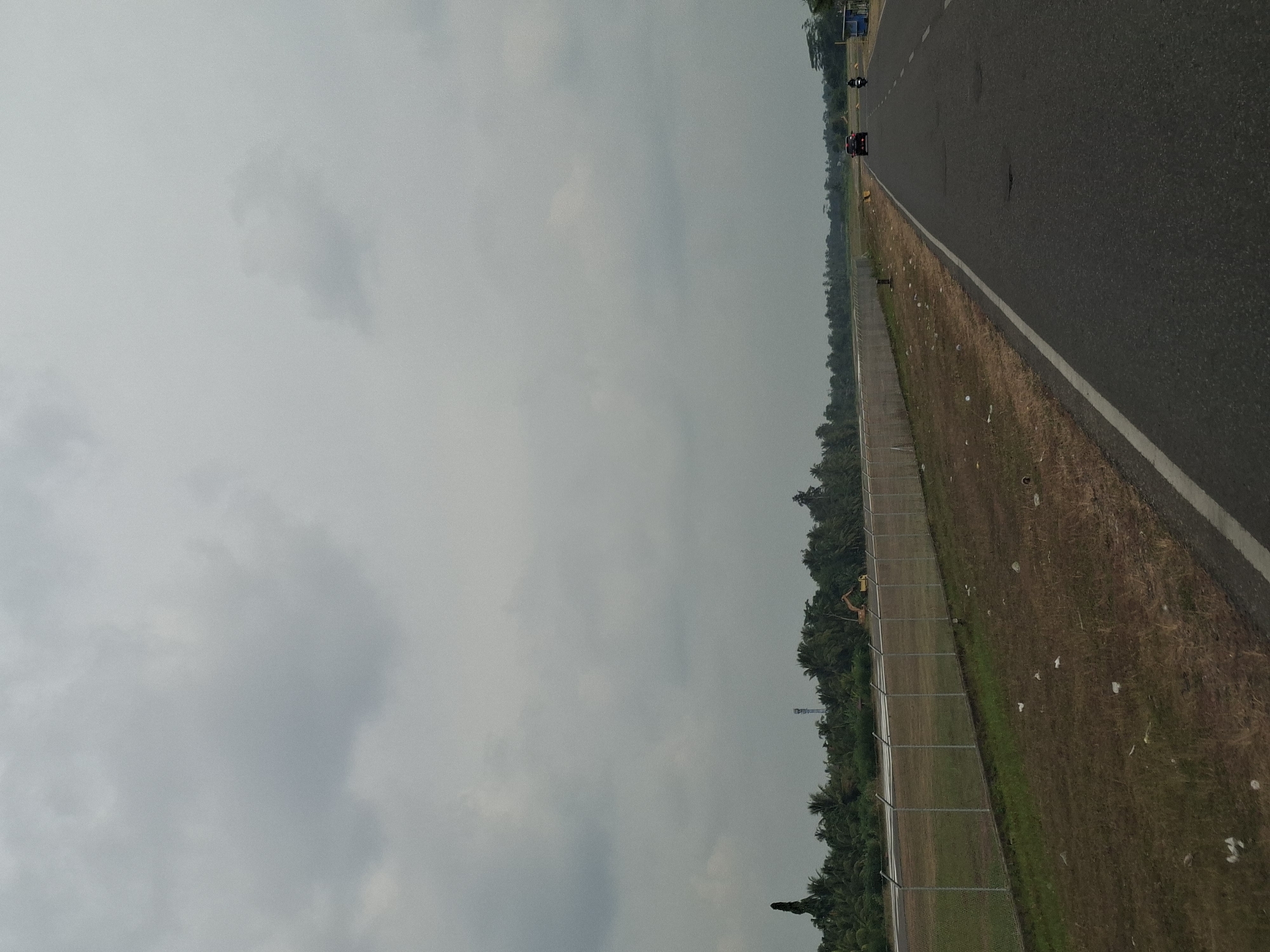 The image depicts a view from a road in Peurupok, Indonesia, with a cloudy sky overhead. To the left of the road, a fenced-off area with dry grass and scattered white debris is visible, beyond which lies a treeline of palm trees and other vegetation. A tall, slender structure, possibly a telecommunications tower, stands among the trees. Further in the distance, a yellow excavator is parked. On the right side of the road, there are two vehicles: a dark-colored car and a motorcycle, both traveling away from the viewer. The road itself is asphalt, with a white lane marker.