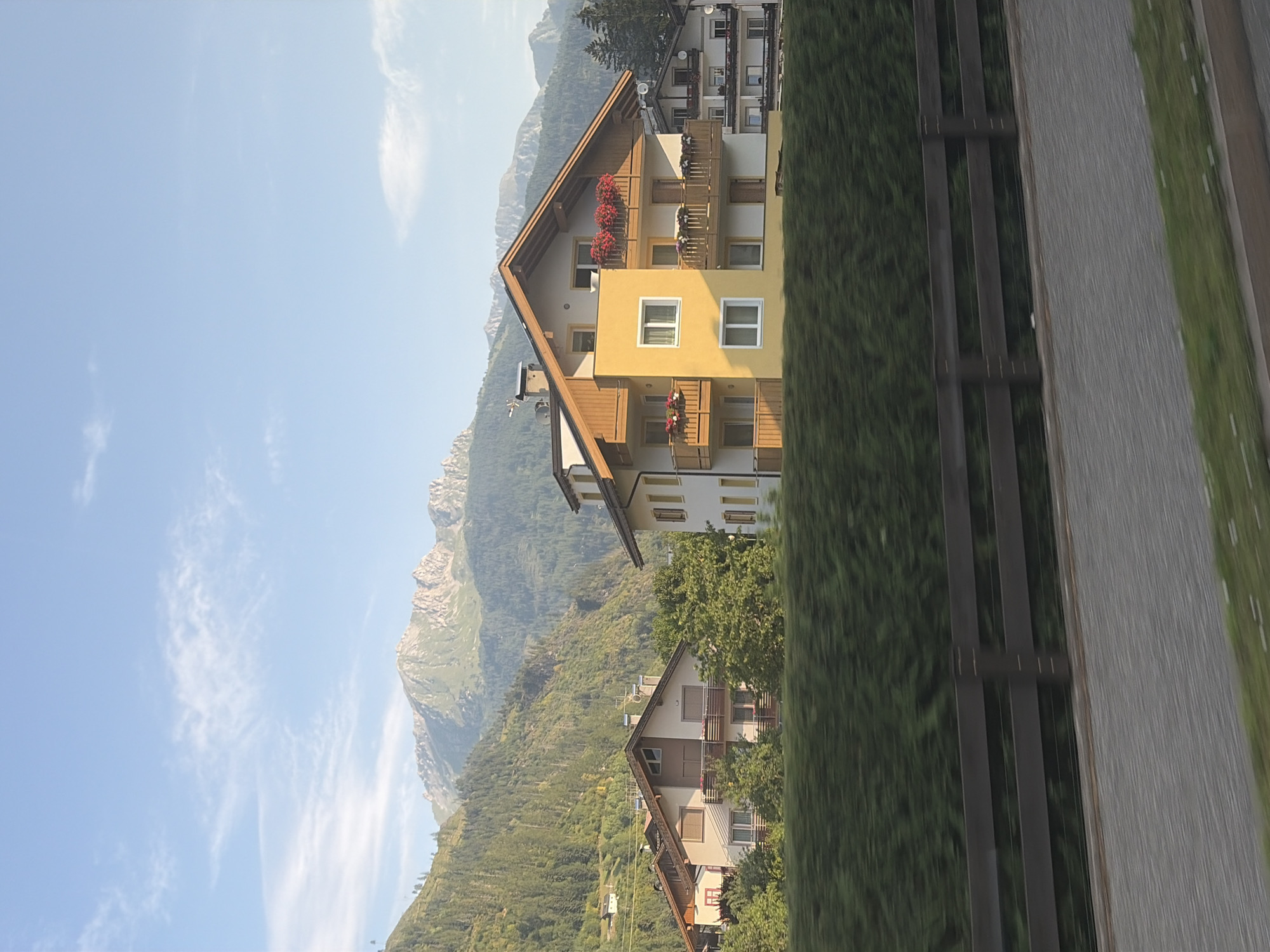 The image depicts a daytime scene in Predazzo, Italy, featuring the iconic Dolomites mountain range in the background. In the foreground, several buildings with a traditional Alpine architectural style are visible, characterized by steep roofs and wooden balconies. One prominent building is painted yellow and white, adorned with red flowers in window boxes. A dense green hedge runs horizontally across the middle ground, partially obscuring a wooden fence and a paved walkway. The sky is a clear blue with scattered white clouds. The overall impression is that of a serene mountain village during daylight hours.