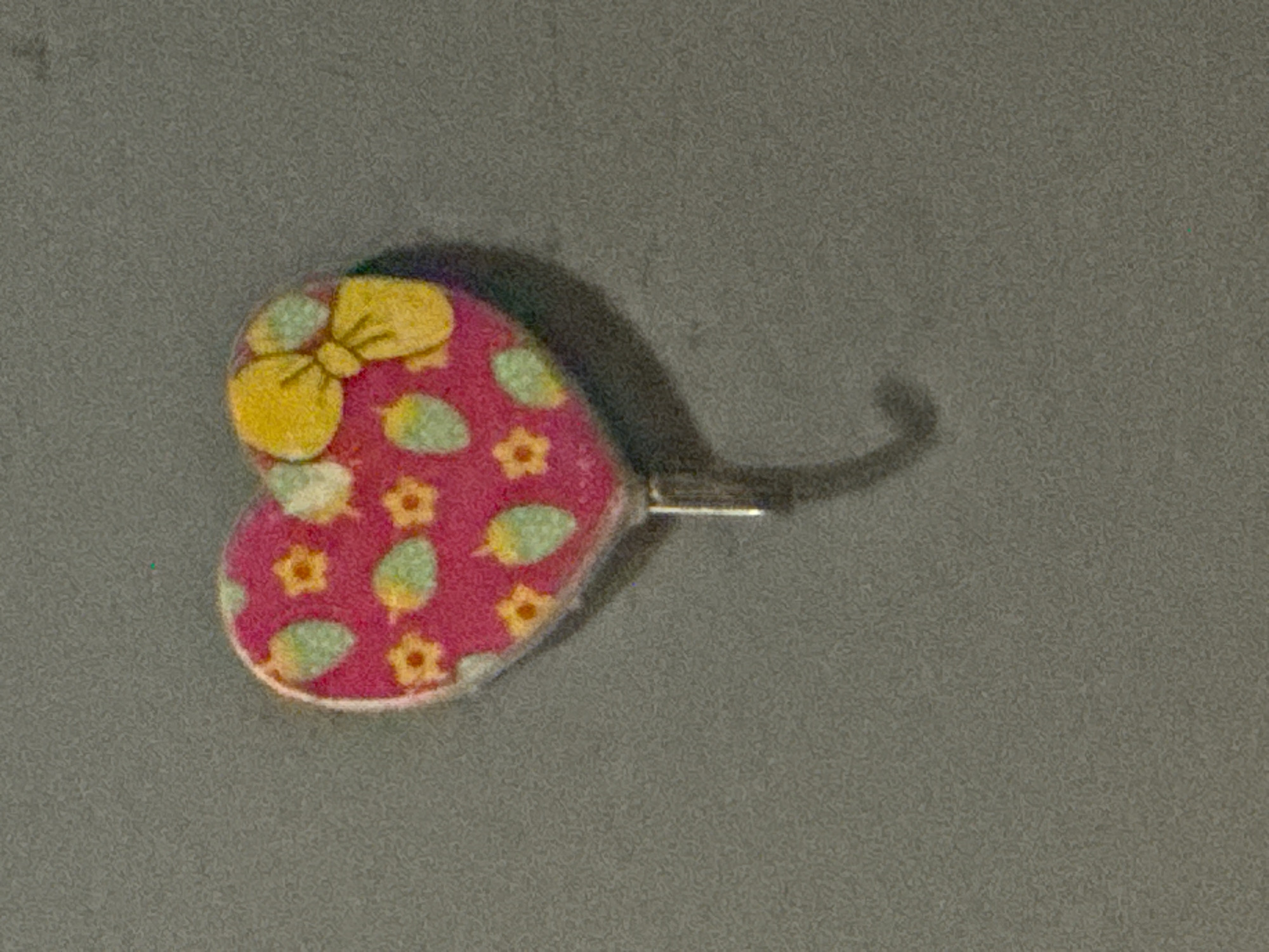 A close-up, still-life photograph features a decorative, heart-shaped pin or brooch laid against a textured, uniform gray background. The heart is primarily pink, adorned with a vibrant yellow bow design on its upper left lobe. The surface of the heart is further decorated with small yellow, five-petal flowers and light green, leaf-like shapes. A thin white border frames the perimeter of the heart. A metallic pin mechanism extends from the back of the heart, casting a shadow to its right, alongside the shadow cast by the object itself, suggesting a light source from the upper left. The gray background exhibits a fine, granular texture. The image shows no human subjects or overt activity, focusing solely on the intricate details of the decorative item. There is no visible text. The scene is set in Kurunduma, Nigeria.