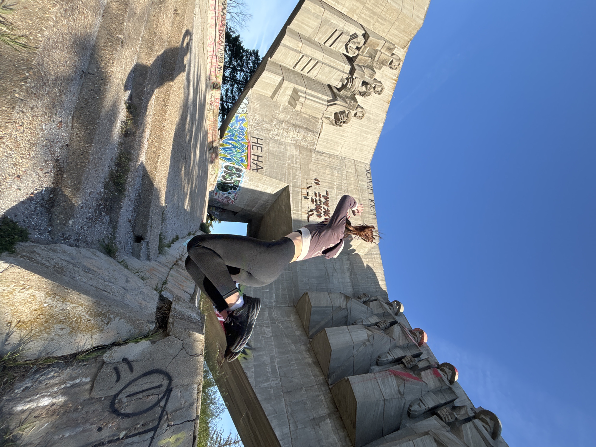 A low-angle, rotated photograph captures a woman in athletic wear jumping or posing on a monumental concrete structure, likely a monument or building, against a bright blue sky. The woman wears a long-sleeved purple top, black leggings, and black sneakers with pink and white striped socks. Her brown hair is loose and flowing, suggesting movement. The structure appears to be an abandoned or decaying monument, heavily graffitied, with a central archway. The concrete is weathered and cracked. The architecture features bas-relief sculptures of figures on the monument's face. The location is likely the Monument of the Bulgarian-Soviet Friendship in Varna, Bulgaria, based on the architectural style and decay. The photograph creates a sense of scale, juxtaposing the human form against the massive monument, and conveys a sense of urban exploration.