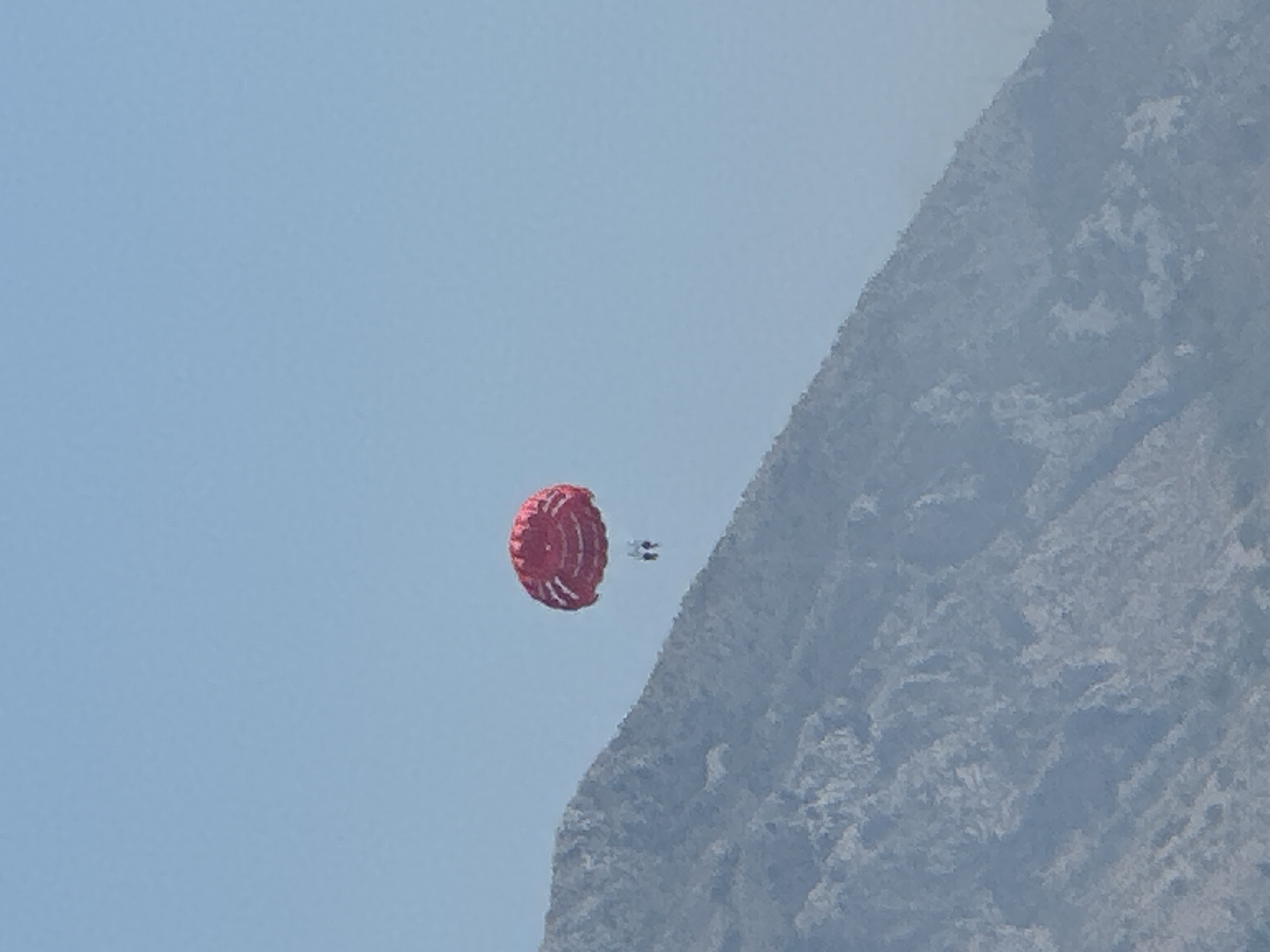 In the city of Boreti, Montenegro, a person is parasailing with a red parachute. The parachute is visible against a clear blue sky. A mountain is in the background, and the person is seen in the distance. The weather appears to be good, with a lot of sunlight.