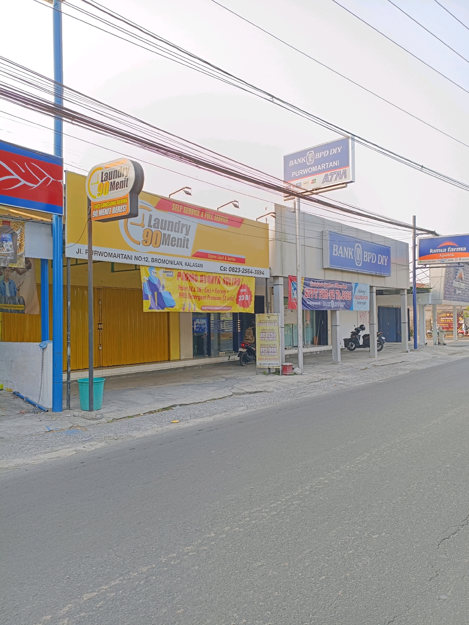 This is a street-level view of businesses in Purwomartani, Indonesia. On the left, a yellow building with a prominent "Laundry 90 Menit" sign is visible, indicating a 90-minute laundry service. The address is listed as Jl. Purwomartani NO.12, Bromonilan, Kalasan. Adjacent to the laundry, another sign with red and blue elements is partially visible. To the right of the laundry, a building with a blue sign reading "BANK BPD DIY PURWOMARTANI ATM" signifies a BPD DIY bank branch with an ATM. Further to the right, a "Kimia Farma Apotek" sign indicates a pharmacy. The street in front of these businesses is paved and shows some wear and tear. Power lines are strung overhead. A green trash can is located on the sidewalk in front of the laundry. Two motorcycles are parked in front of the bank. The overall scene depicts a commercial strip in a town.