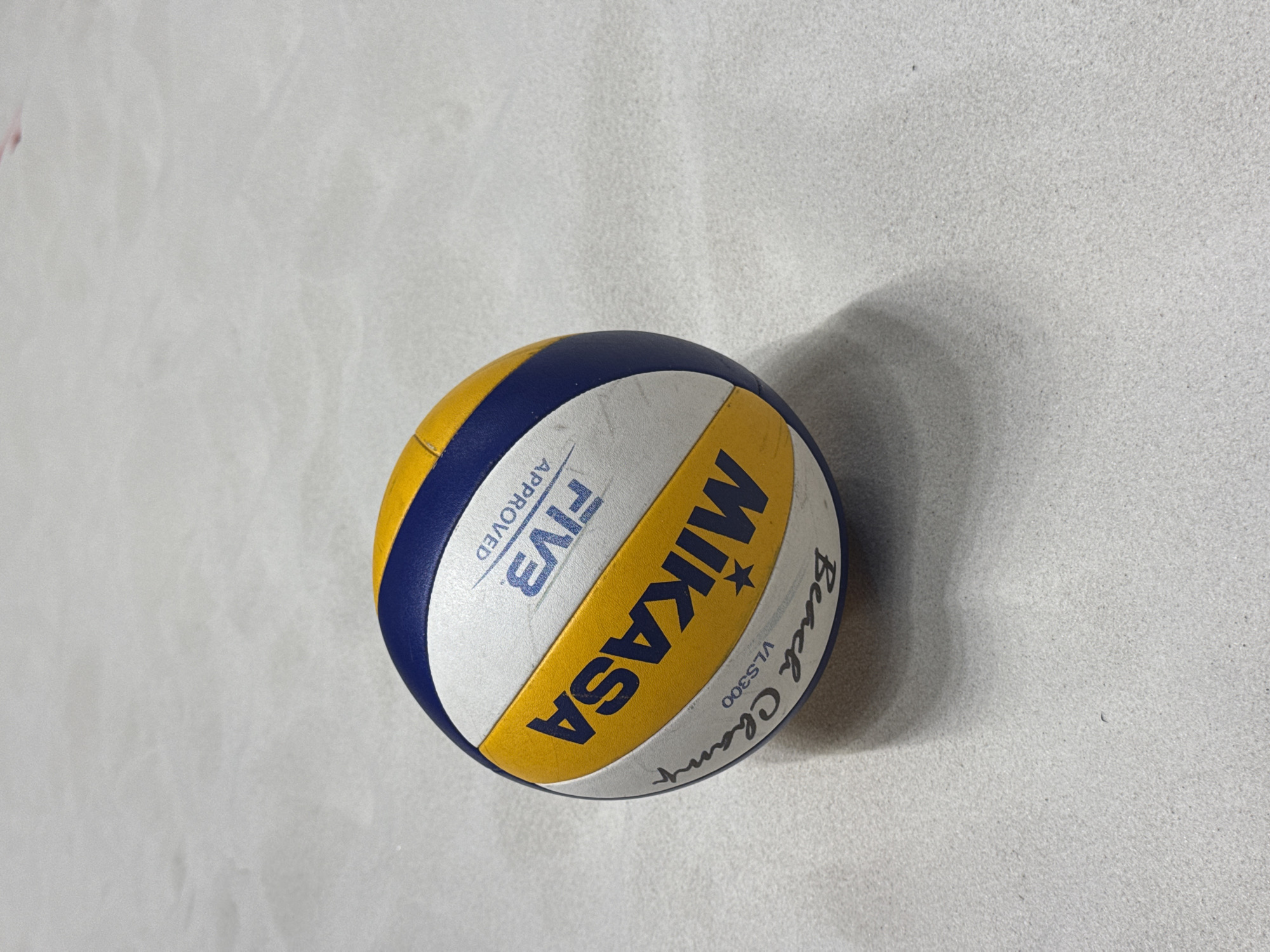 A Mikasa volleyball, predominantly blue, yellow, and white, is the central subject of this image, resting on a light grey, textured surface. The ball is positioned slightly off-center towards the right. Its panels alternate in color, featuring dark blue and vibrant yellow sections, separated by white strips.
Clearly visible text on the ball includes the brand "MIKASA" in bold yellow letters, accompanied by a star logo. On an adjacent white panel, "FIVB APPROVED" is printed vertically. Further around the ball, in a cursive style, "Beach Champ" is written, with "VLS300" directly beneath it. The ball shows signs of moderate use, with some scuff marks and dirt visible on its surface, particularly on the white and yellow panels, indicating it has been played with.
The background is a uniformly lit, light grey surface that appears to be concrete or a similar fine-grained flooring material. The texture is subtle but discernible. A soft, diffused shadow is cast by the ball towards the right, suggesting indirect or ambient lighting. There are no people visible, and no activity is occurring; the ball simply rests on the ground. The scene is devoid of specific location cues, though the context provided indicates the location is Krasnoyarsk, Russia. The image is a close-up, focusing entirely on the ball and its immediate surroundings.