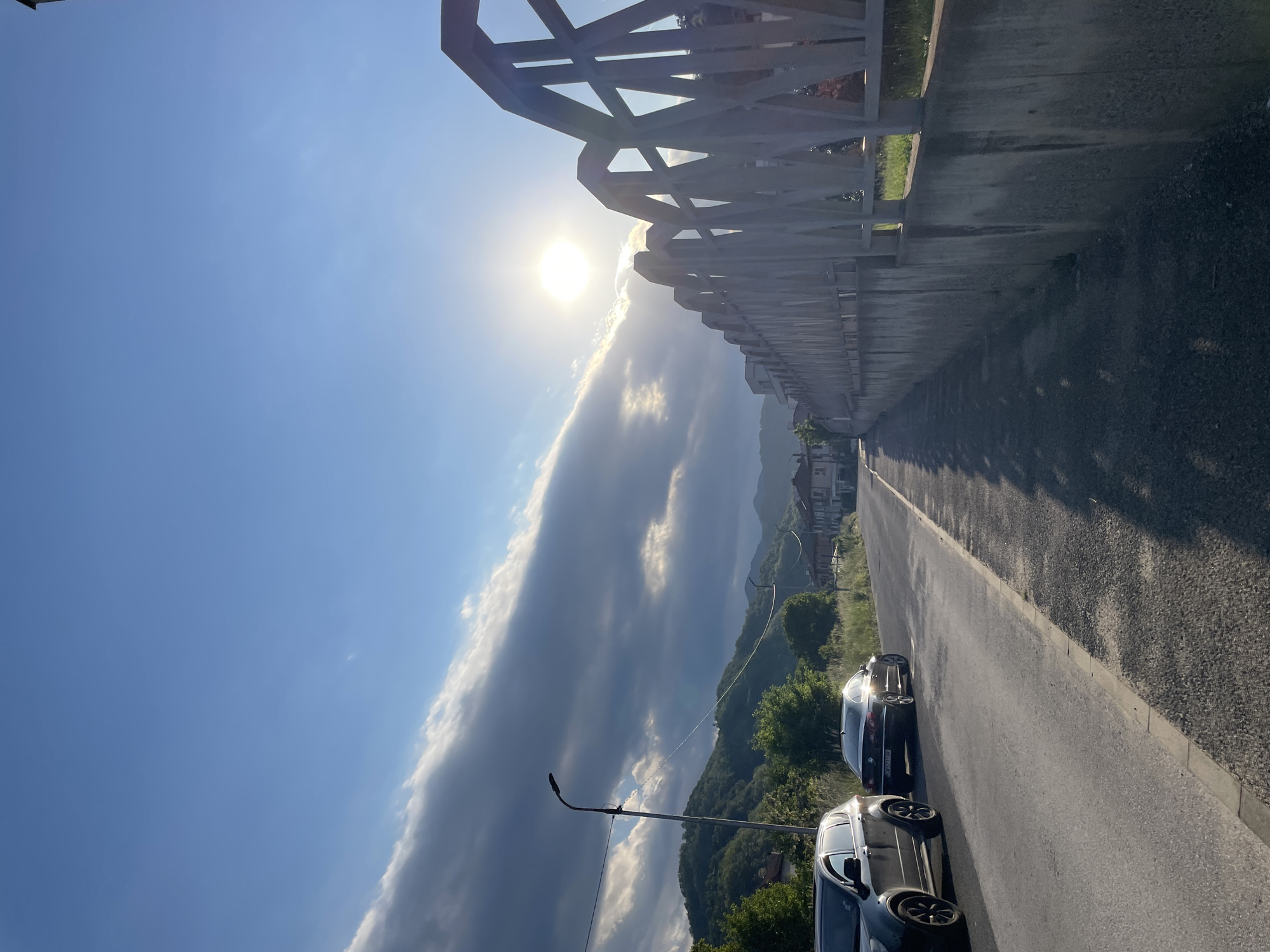 The image shows a road in Zlatograd, Bulgaria, with two cars parked on the side. A metal fence runs along the roadside. In the background, there are rolling hills with scattered houses and trees. The sky is blue with a long band of white and gray clouds, and the sun is shining brightly. A street lamp is visible on the left side of the road.