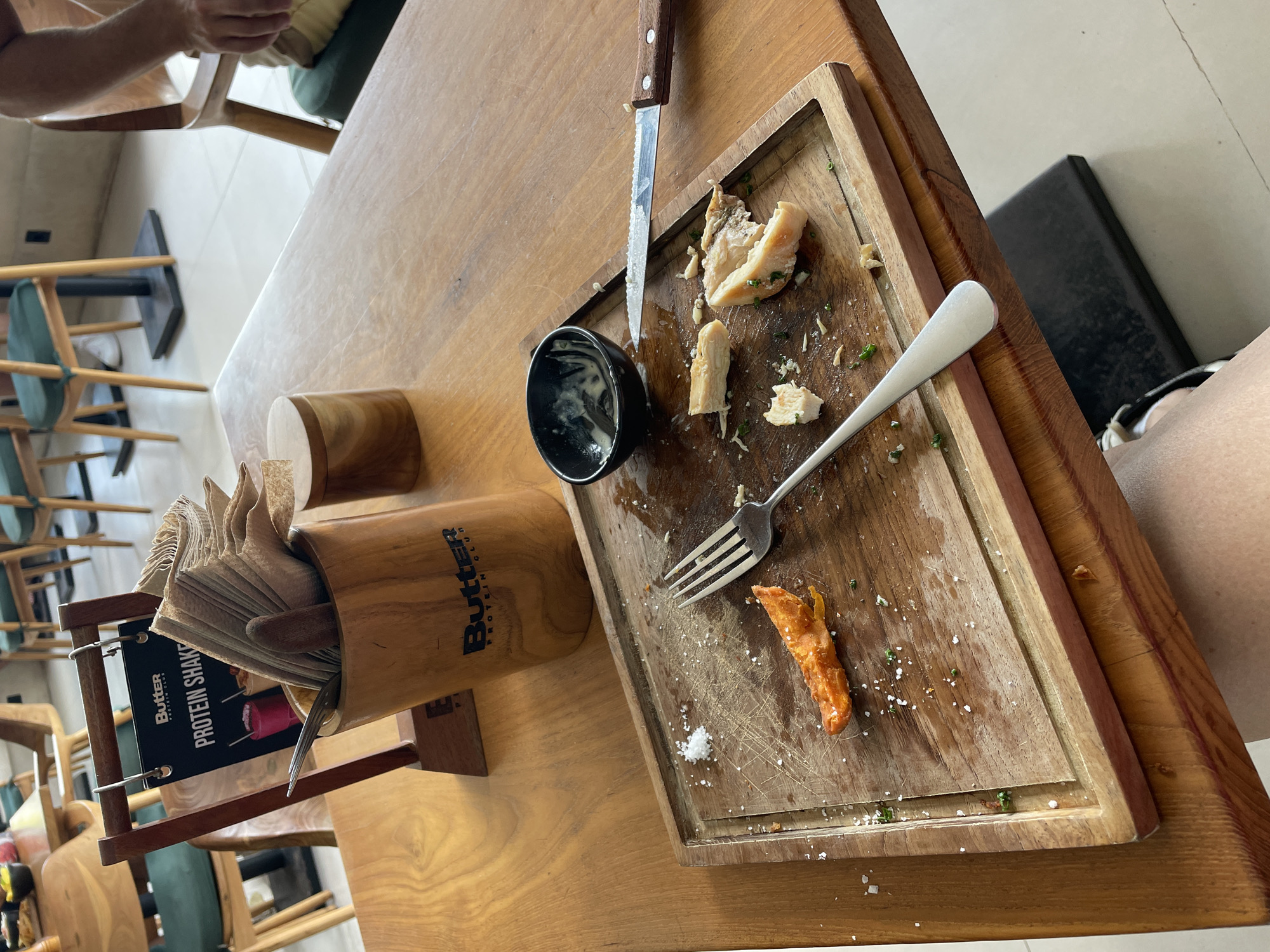 The overhead, slightly angled shot captures a wooden table at a restaurant in Mengwi, Indonesia, with the remnants of a meal. The table is set with a wooden board that holds a single, partially eaten piece of orange food, likely a sweet potato wedge, and a small pile of salt crystals. Scattered across the board are crumbs and some chopped green herbs. A silver fork rests on the board, pointing towards the bottom left, and a serrated knife with a wooden handle lies to the right. A small, black bowl with a white creamy sauce sits at the top center of the board.
In the background, a wooden napkin holder stands, filled with folded paper napkins. Behind it, a wooden menu stand displays a "Butter Protein Shake" menu. Various chairs with green cushions are visible in the restaurant setting, and a person's hand and arm, wearing a light-colored sleeve, are seen resting on a chair to the right of the frame. The overall impression is of a recently concluded meal in a casual dining environment.