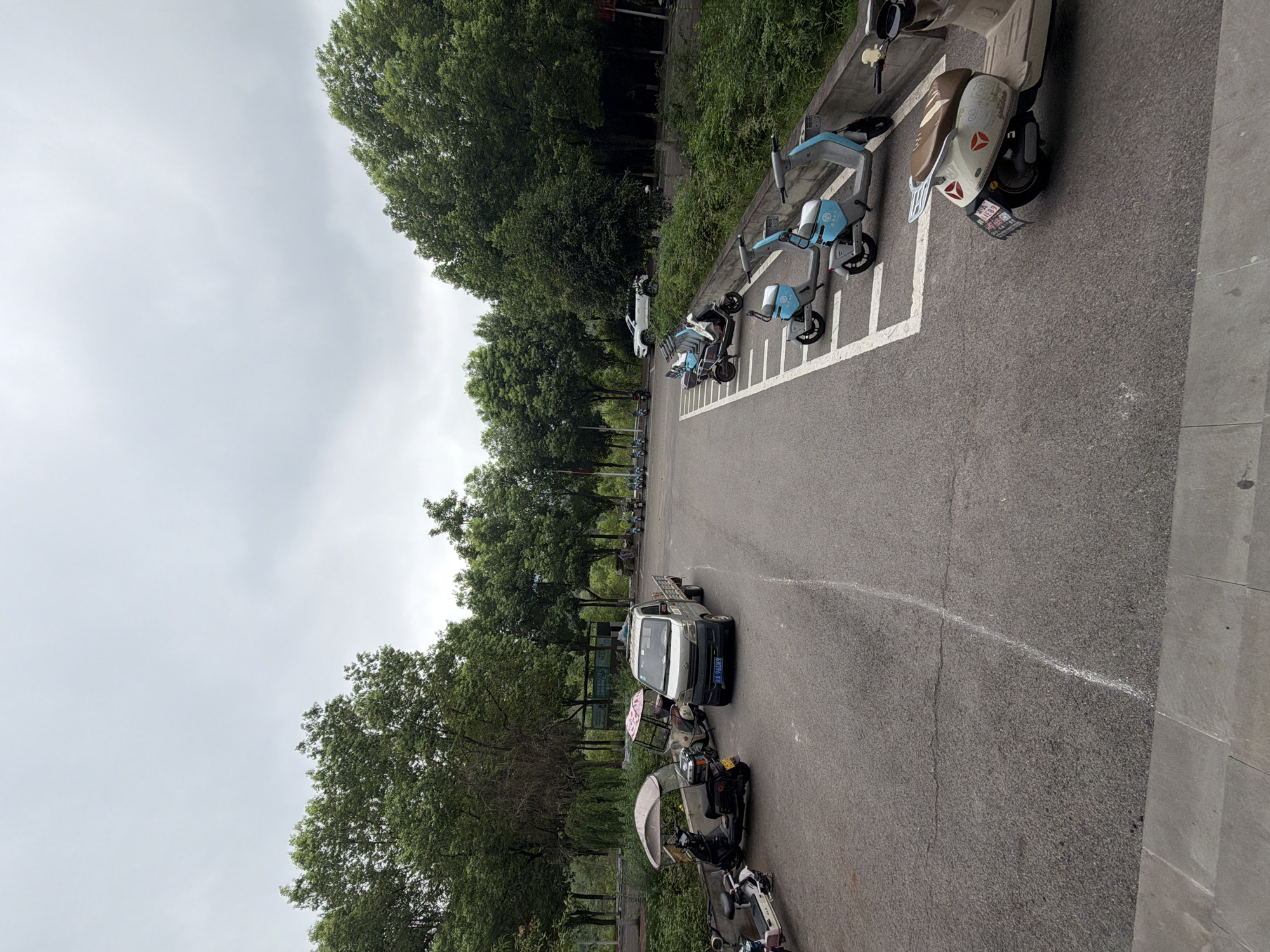 This is a medium-shot, eye-level, outdoor photograph taken on a cloudy day. The scene depicts a parking area for various vehicles, including electric scooters and a small truck, located in the city of Jiangjin, China. In the foreground, a light-colored electric scooter with a brown seat is parked facing right. To its left, a row of light blue and white electric scooters are parked in designated white-lined spaces, facing left. Beyond these, several other electric scooters and a small silver truck with a license plate reading "A 96DK9" are parked. In the background, lush green trees line the perimeter of the parking area, with a dense forest visible further back. A white SUV is parked in front of the trees on the right side of the frame. The ground is asphalt, with white lines marking parking spaces. A concrete curb is visible along the right side of the parking area.