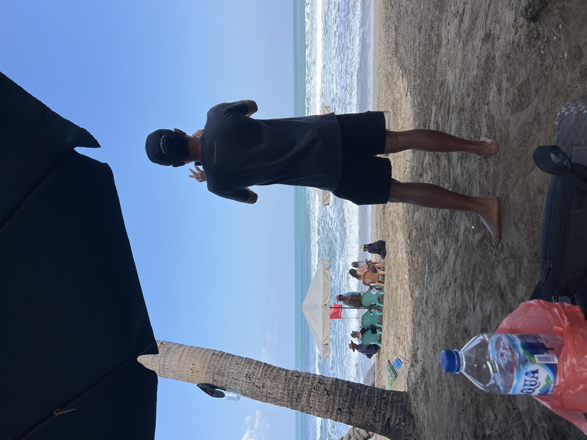 A man stands on a sandy beach in Kuta, Indonesia, facing the ocean and holding up a phone to take a picture. He is wearing a dark t-shirt and black shorts. To his left, a large palm tree trunk is visible, and a dark umbrella is overhead, casting shade. In the background, several people are sitting on chairs under a light-colored umbrella, facing the water. The ocean has moderate waves, and the sky is clear and blue. In the foreground, a plastic bottle of water and a red plastic bag are on the sand.