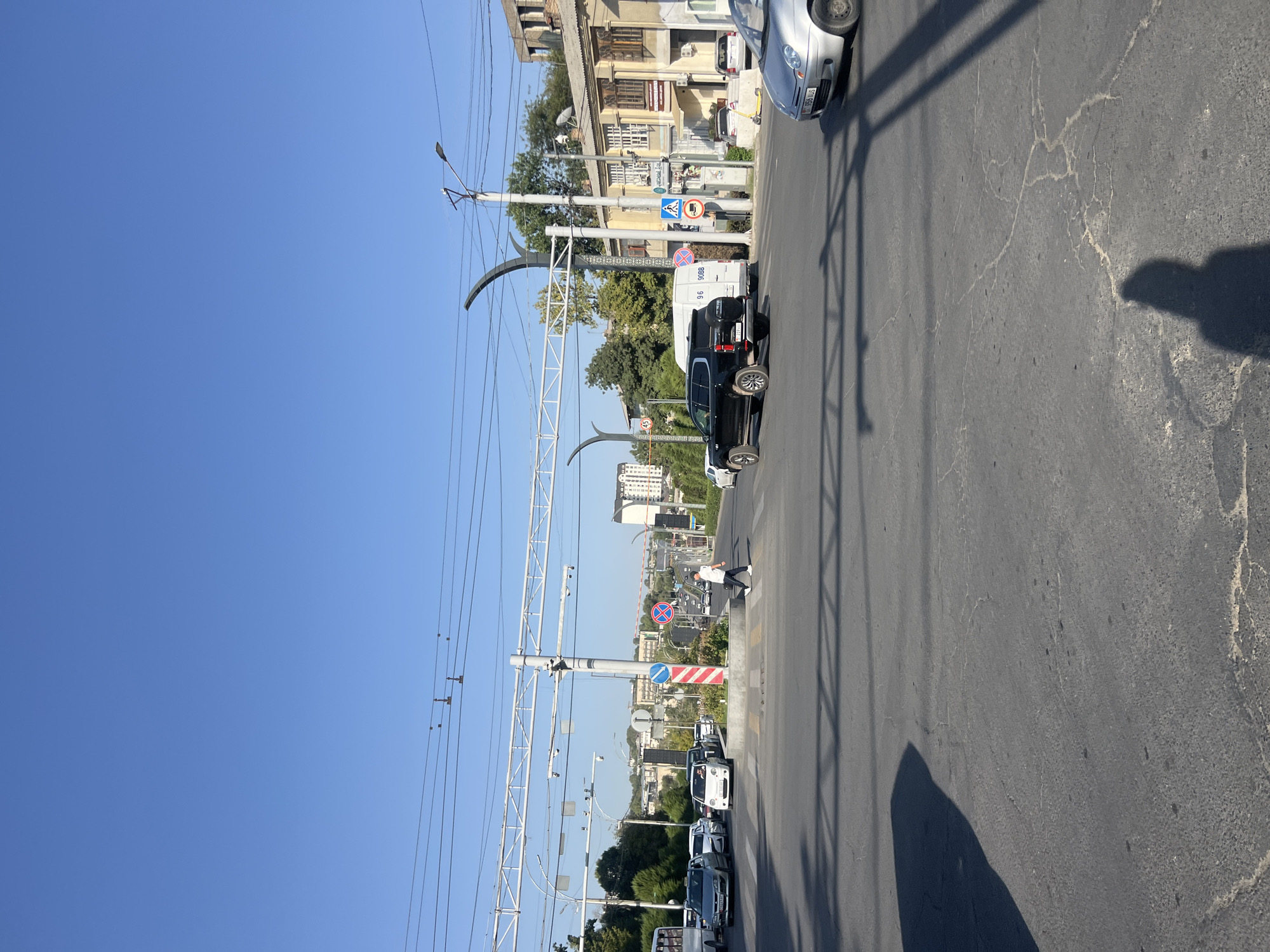 A street-level view captures a sunlit intersection in Osh City, Kyrgyzstan, on a clear day with a vibrant blue sky. The foreground is dominated by asphalt pavement, marked with faded white lines and illuminated by the shadow of a person and a triangular structure. A silver sedan is partially visible in the lower right quadrant of the image.
The intersection features multiple lanes of traffic and a pedestrian crossing. Several vehicles are present, including a black SUV in the center of the frame, a white van, a white compact car, and several other cars and a bus further in the background. A pedestrian is crossing the street from left to right, silhouetted against the sunlit road.
Traffic signals and overhead electrical wires are visible, along with streetlights and poles. A concrete median with some foliage separates parts of the intersection. Buildings with traditional architecture and modern structures can be seen in the background, with trees and vegetation interspersed. The overall scene depicts a typical urban traffic environment with clear visibility and moderate activity.