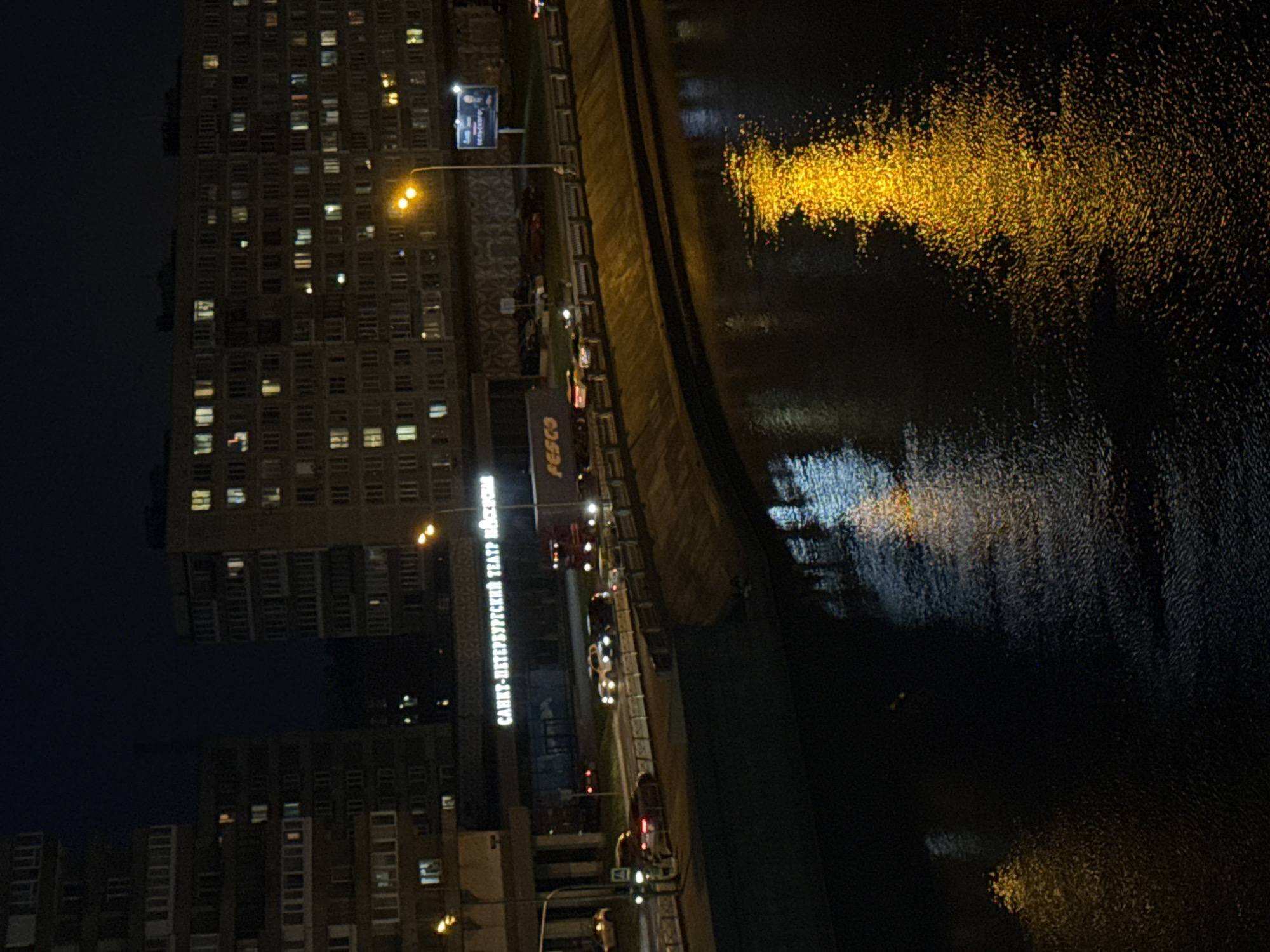 This nighttime image, captured from a high vantage point, depicts an urban scene in Saint Petersburg, Russia, overlooking a river or canal alongside a street and buildings. The sky is dark, indicating late evening or night.
To the left, a large, multi-story building dominates the skyline, its numerous rectangular windows glowing with warm light, suggesting occupied apartments or offices. Below and to the right of this building, a street or embankment runs parallel to the water. Several vehicles, including cars and possibly a red truck, are visible on the road, with their headlights and taillights creating streaks of light. Streetlights emit a bright, warm yellow glow, illuminating sections of the pavement and contributing to the reflections on the water.
The body of water, likely a river or canal, occupies the right side of the frame. Its dark surface is intensely reflective, particularly catching the vibrant, scattered golden-yellow light from the streetlights and potentially nearby buildings. This creates a striking shimmer across the water, interspersed with fainter white and blue reflections.
Visible text on a prominent sign on a building confirms the location, reading "САНКТ-ПЕТЕРБУРГСКИЙ ТЕАТР МЮЗИКЛА" (Saint Petersburg Musical Theater). Another sign below it displays "FESCO". A billboard further down the street shows text that includes "БЕЛУГИН" or similar fragmented words. The overall scene captures the quiet, illuminated activity of a city after dark.
