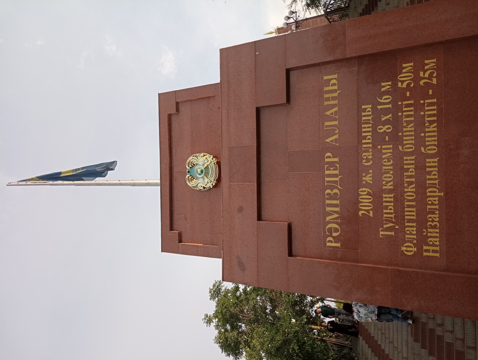 This media file is a vertical shot of a monument with inscriptions in Kazakh. The monument is made of red granite and features a golden emblem, likely a national symbol. The inscriptions, written in large golden letters, provide information about the monument, indicating it was built in 2009, its dimensions (flag size 8x16m, flagpole height 50m, and spear height 25m). The monument appears to be situated in a public space, possibly a plaza or park, with a flag pole flying a Kazakh flag visible to the left. Some people are seen standing on stairs in the lower right portion of the image, and trees are present in the background. The sky is bright, suggesting daylight. The overall scene depicts a landmark with historical or civic significance in Shymkent, Kazakhstan.