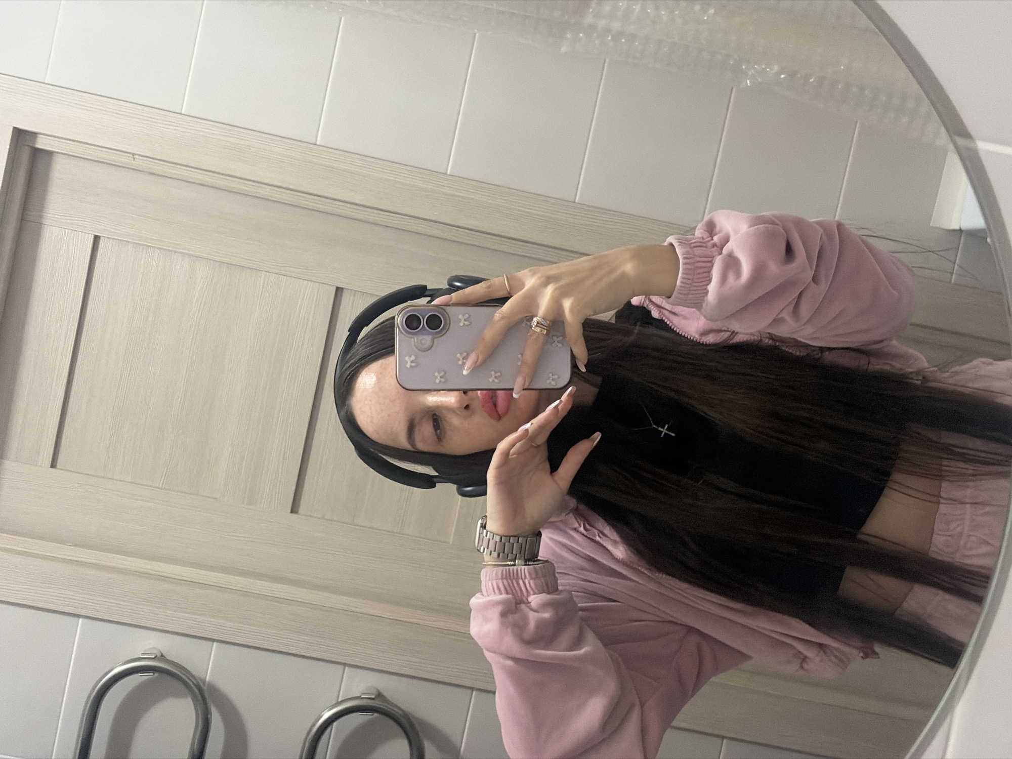 A young woman with long dark hair wearing headphones and a pink tracksuit is taking a selfie in a mirror. She is holding her phone with her left hand. She is wearing a silver watch on her left wrist and rings on the fingers of her left hand. The phone has a lavender-colored case decorated with white flowers. She has a cross necklace. The reflection in the mirror shows a bathroom with white tiled walls and a light wood-paneled door. A towel rack with metal handles is visible below the mirror. The location is Saint Petersburg, Russia.