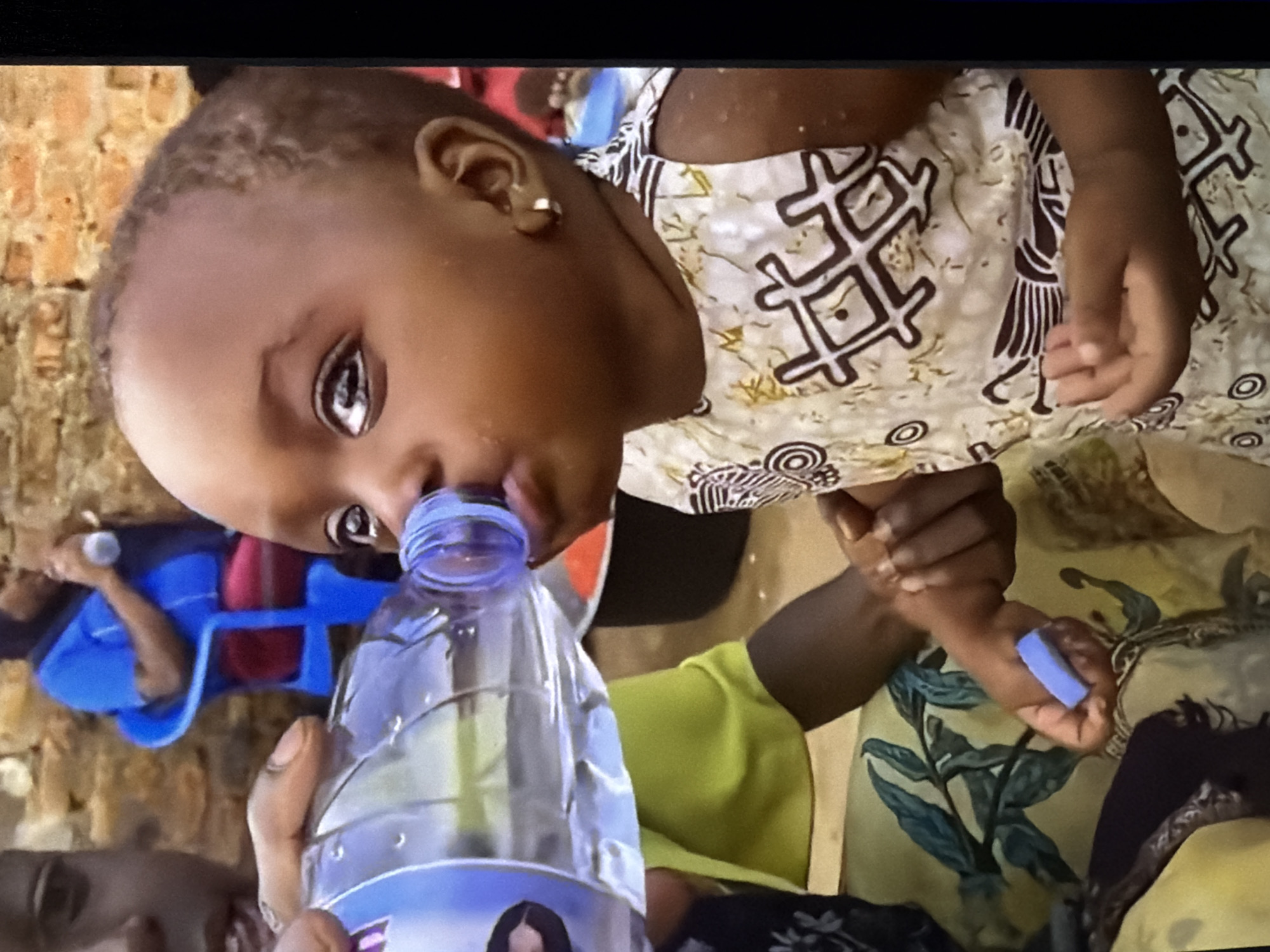 A close-up, eye-level shot shows a young child drinking from a water bottle in an outdoor, arid environment. The child, positioned slightly right of center, has dark skin and wears a light-colored dress with a dark, geometric pattern. Their left eye is prominently featured, wide open and looking towards the bottle. A bottle of water, held by an unseen person's hand on the left, is tilted to the child's mouth. Behind the child, a person in a blue shirt and red pants sits on a blue chair, holding a similar bottle. The background consists of a brick wall and a textured surface, suggesting an urban or semi-urban setting.