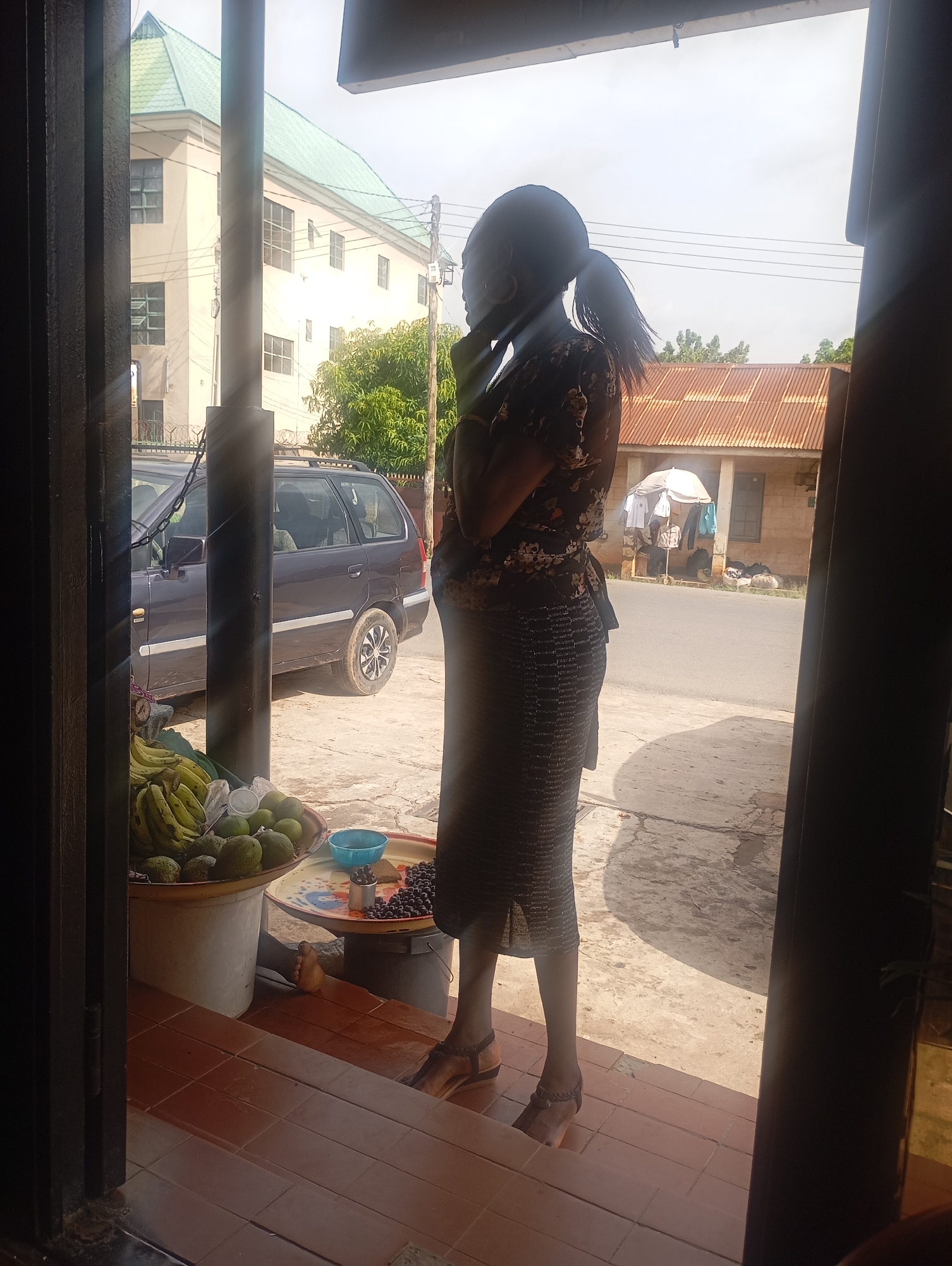 A woman with her hair in a ponytail stands by a doorway, looking out at a street scene. She is wearing a patterned blouse and a long, patterned skirt, and her sandals are visible. In the foreground, on a tiled step, are two round trays. One contains a pile of green fruits, possibly avocados, and a bunch of bananas. The other tray holds a blue bowl, some dark berries, and a small container. Behind the woman, a dark colored station wagon is parked on the side of a paved road. Across the street, a building with a corrugated iron roof is visible, with laundry hanging on a clothesline in front of it. Sunlight streams into the scene, casting strong shadows, suggesting it is a sunny day, likely morning or afternoon. The overall setting appears to be a marketplace or residential area in Jos, Nigeria. The woman's posture and gaze suggest she is observing the activity outside.