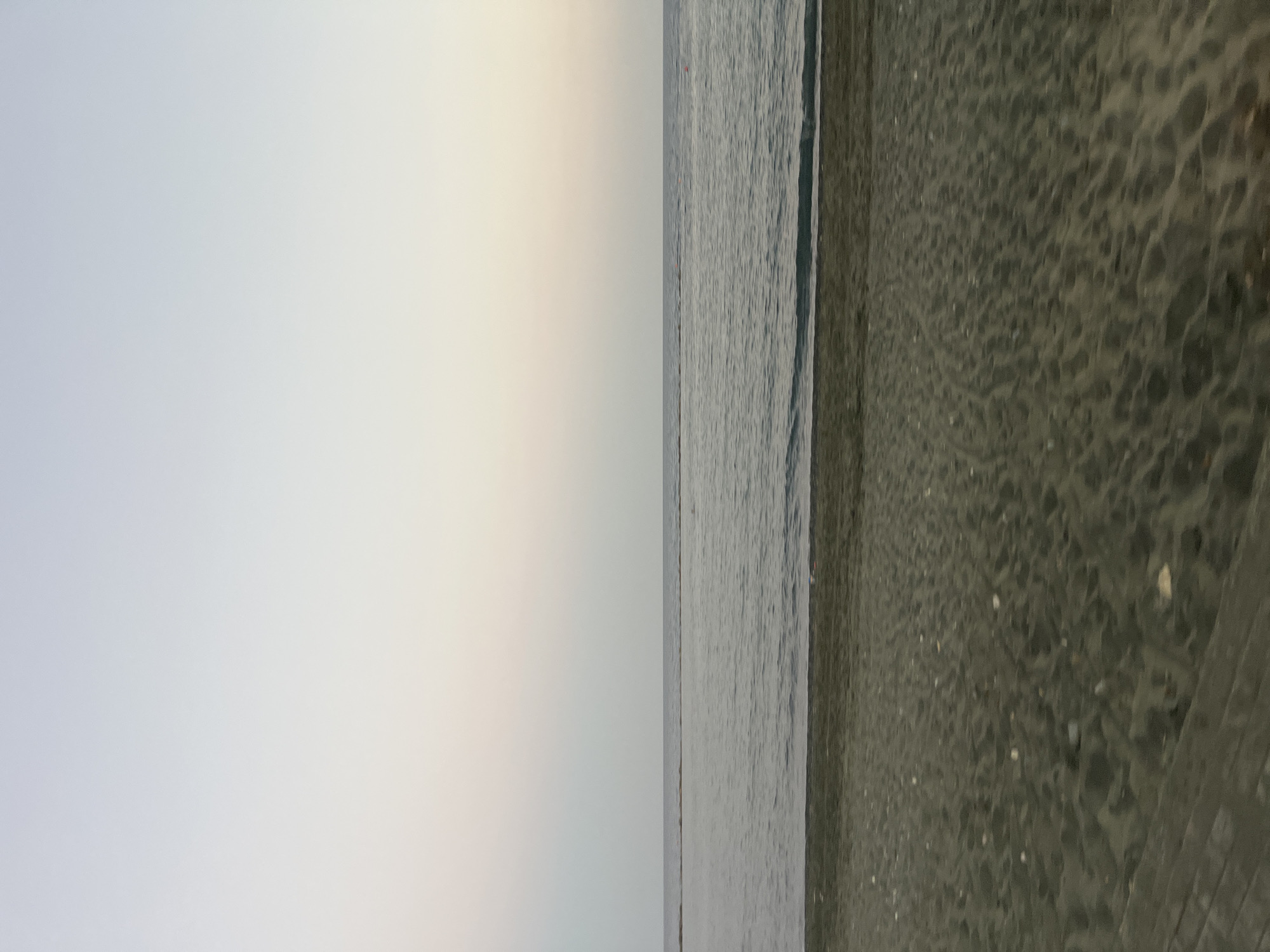 The image shows a wide, horizontal view of a beach and the sea at Agios Tychona Community, Cyprus. The foreground is dominated by dark, textured sand, with small pebbles scattered across its surface. A faint wooden structure is visible on the left edge of the sand. The middle ground features the calm, grey-blue sea, with gentle waves lapping at the shore. A thin, horizontal line, possibly a breakwater or a distant sandbank, stretches across the water. The background is a pale, serene sky, transitioning from a light grey at the horizon to a softer, almost pastel hue at the top. There are no people or discernible objects in the water or on the sand, creating a peaceful, unobstructed coastal landscape.